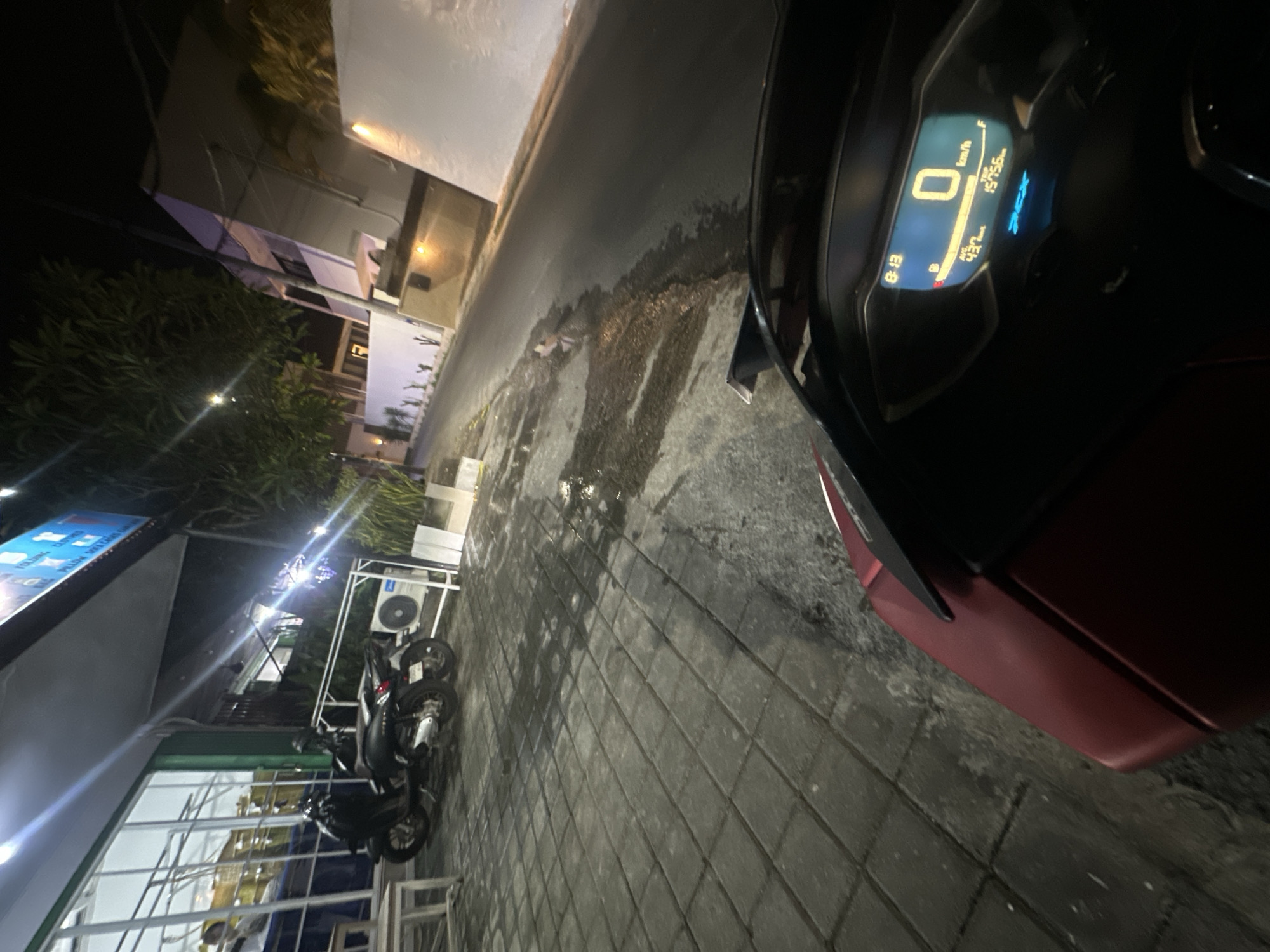 The image presents a nighttime scene in North Kuta, Indonesia. The foreground is dominated by the dashboard of a stationary scooter, showing a speedometer reading of 0 km/h and an odometer reading of 15756 km. The time displayed is 8:13. In the background, two scooters are parked outside a storefront. The building appears to have a sign with blue text. A white air conditioning unit is visible mounted on a metal frame near the parked scooters. The scene is lit by streetlights and the illumination from the building. Pavement is visible, reflecting the ambient light.