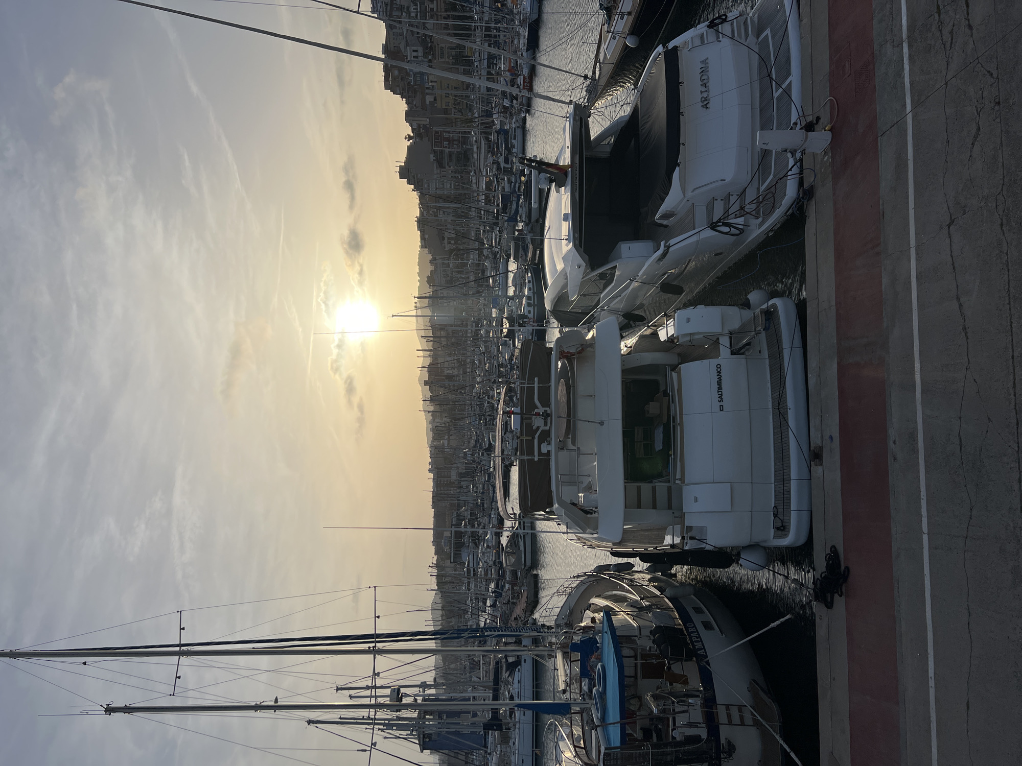 The image depicts a marina filled with numerous yachts and sailboats at sunset. The location is likely in El Masnou, Spain, a coastal town known for its harbor. The sun is low in the sky, casting a golden light over the scene. The harbor is densely populated with boats of varying sizes, their masts creating a network of lines against the sky. Buildings are visible in the background, suggesting an urban setting near the marina. Three yachts are docked prominently to the right in the foreground of the image.