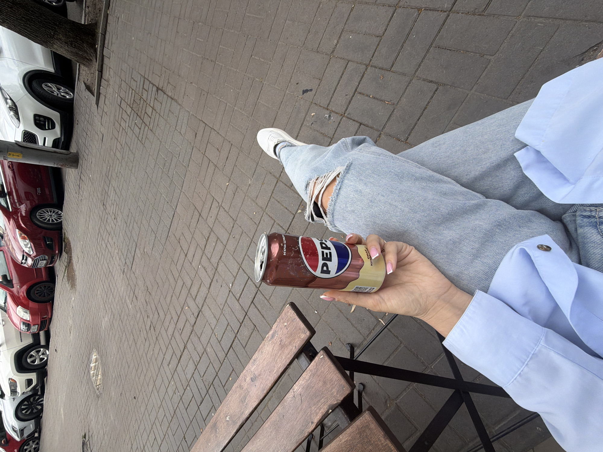 A person is holding a can of Pepsi while sitting on a park bench in Kyiv, Ukraine. The person is wearing ripped jeans and a light blue button-down shirt. The background shows a sidewalk paved with brick-like stones, with several cars parked along the street. A manhole cover is visible on the sidewalk in the distance. The overall scene is casual and relaxed.