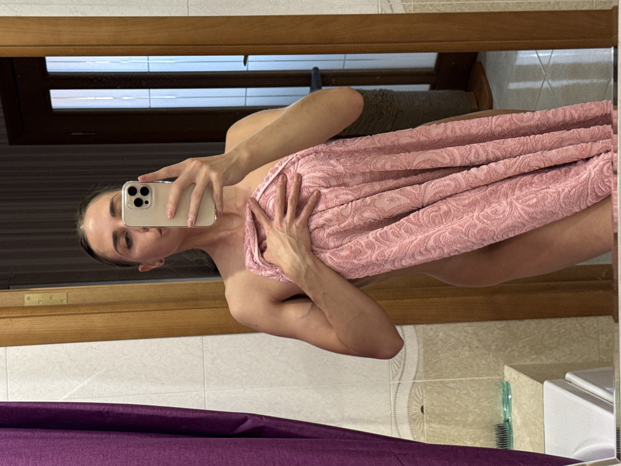 This image is a mirror selfie taken by a person in what appears to be a bathroom, with the photograph rotated 90 degrees counter-clockwise.
**Subjects:** The central subject is a person, likely female, visible from the chest upwards, holding a light-colored smartphone with a multi-lens camera module directly in front of their face. Their expression is neutral, looking towards the phone's camera. The person is wrapped in a textured, pink towel with a subtle pattern.
**Objects and Scene:** The entire scene is a reflection in a mirror. The background shows light-colored tiled walls, some with decorative patterns. A wooden frame surrounds the mirror. At the bottom of the image, a section of purple fabric, possibly a shower curtain or bath mat, is visible. On the right, parts of a white ceramic fixture (like a toilet or sink) and a green comb or brush resting on a counter are reflected. A dark, indistinct object is reflected through what might be a window or another mirror at the top.
**Activity:** The person is actively taking a mirror selfie with their smartphone, seemingly after or before a bath, indicated by the towel.
**Notable details:** The primary visual detail is the image's rotation, presenting the person horizontally. No specific time of day or weather is discernible. The setting is clearly an indoor bathroom. No visible text is present. While the location context is given as "Unknown, Russia", this is not visually identifiable from the image itself.