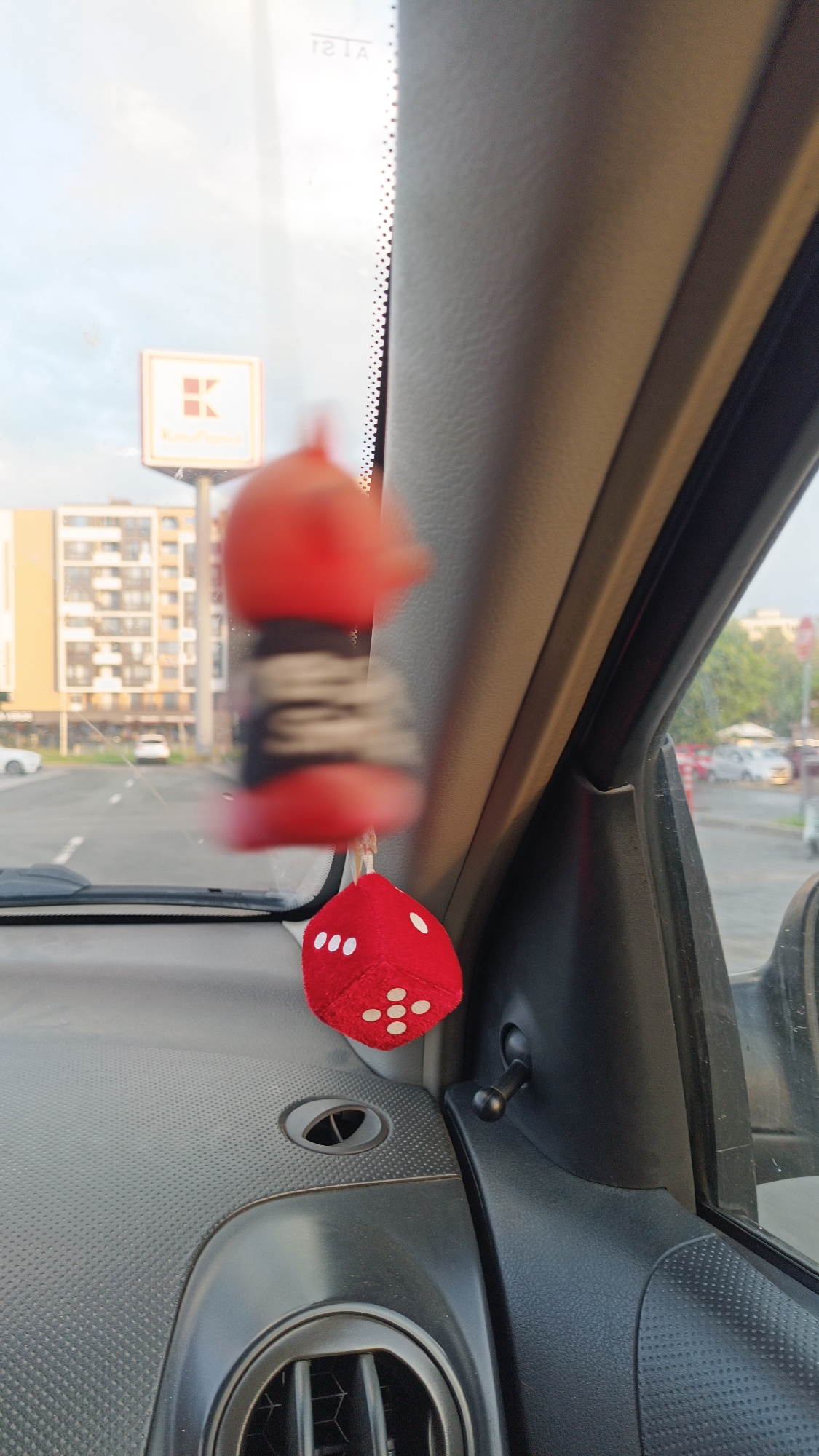 The image shows an interior view from the passenger seat of a car, looking out the front windshield. A red, fuzzy dice with white dots hangs from the rearview mirror. Visible through the windshield is a street scene in Burgas, Bulgaria, with a large Kaufland sign on a building in the background. Other buildings and cars are also visible in the background street scene. The car's dashboard, air vent, and side window are visible in the foreground.