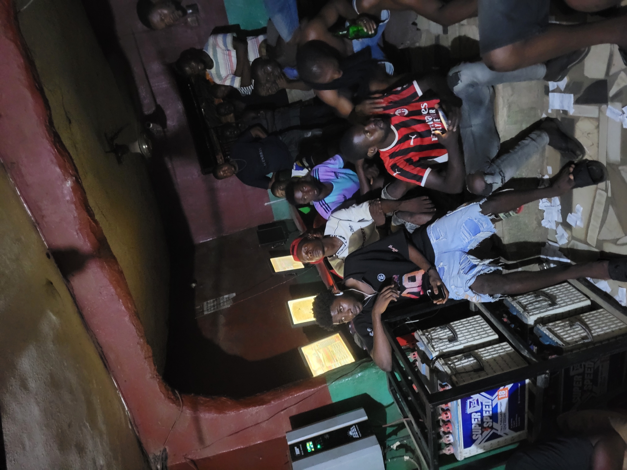 The scene depicts a group of approximately 10 men indoors. They are sprawled on the floor, seemingly relaxing, with some holding what appear to be bottles. The room appears to be dimly lit, with a textured, possibly painted wall visible. Near the bottom of the frame, there's a structure that contains batteries. Given the context, it is likely that the location is within Nkpor, Nigeria.