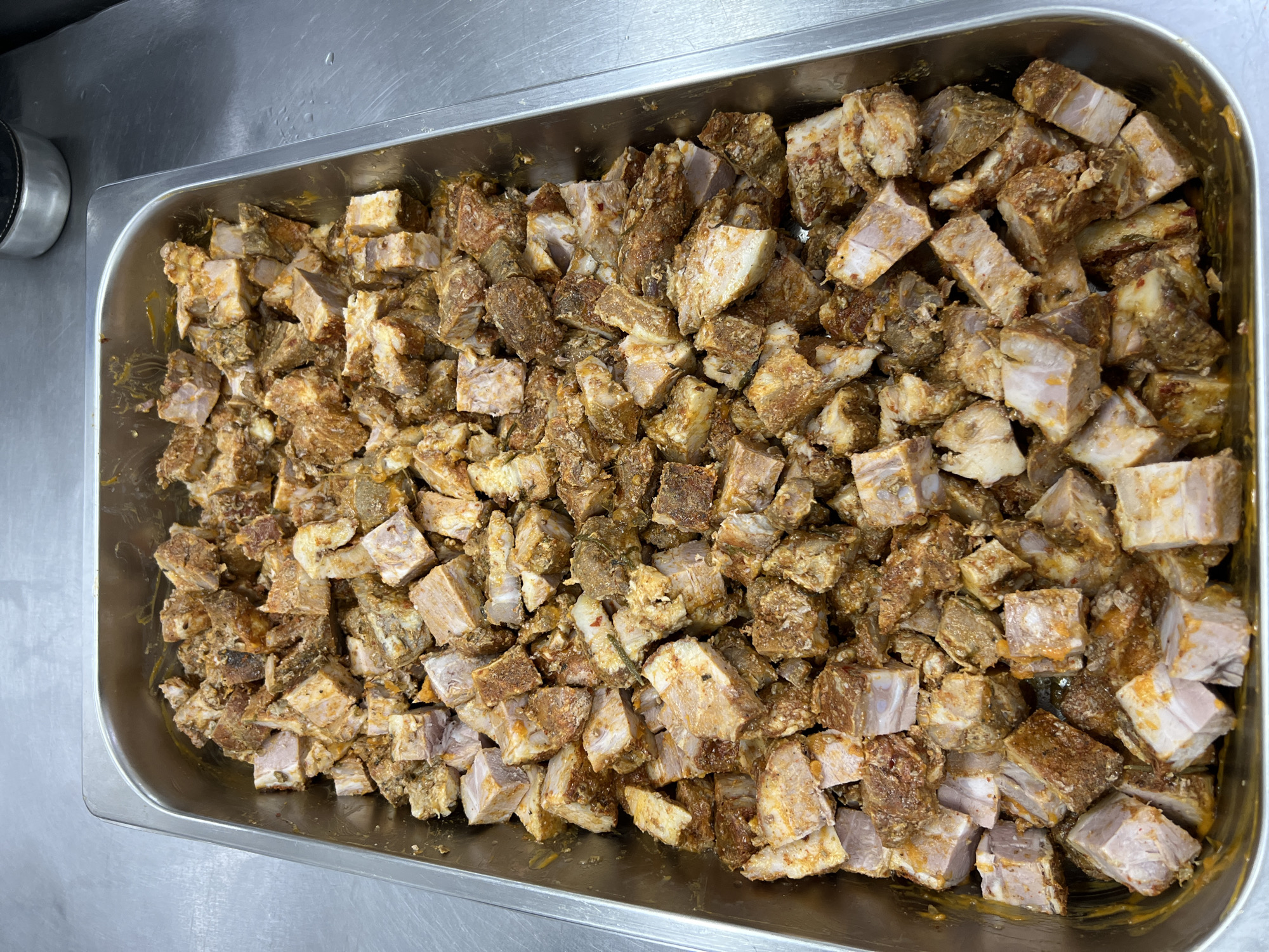 This is a top-down, medium shot of a rectangular metal pan filled with cubed, seasoned pork belly. The pork is cut into roughly one-inch cubes and appears to be raw or unmarinated, with visible layers of meat and fat. The seasoning is a mix of reddish-brown spices, some of which appear to be chili flakes, and possibly herbs, giving the pork a textured, slightly oily appearance. The pan is situated on a stainless steel countertop, likely in a commercial kitchen environment. The lighting is overhead and neutral. There are no people or other objects of significance in the frame. The background is dominated by the metallic surface of the countertop. The location is not specified in the image or context provided.
