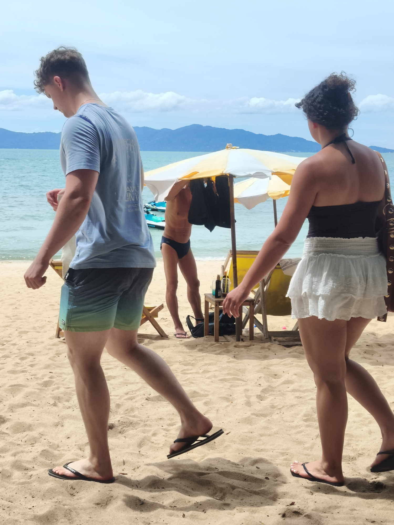 Two people are walking on a sandy beach in Baan Mae Nam, Thailand. In the foreground, a person wearing a blue t-shirt and grey and green patterned swim trunks walks towards the left. Behind them, a person wearing a dark top and a white ruffled skirt walks towards the right. In the middle ground, another person is partially visible, wearing black swim briefs and standing under a yellow and white beach umbrella. Several beach chairs are visible, along with a small wooden table with bottles on it. The ocean is in the background, with distant mountains visible. The sky is partly cloudy.