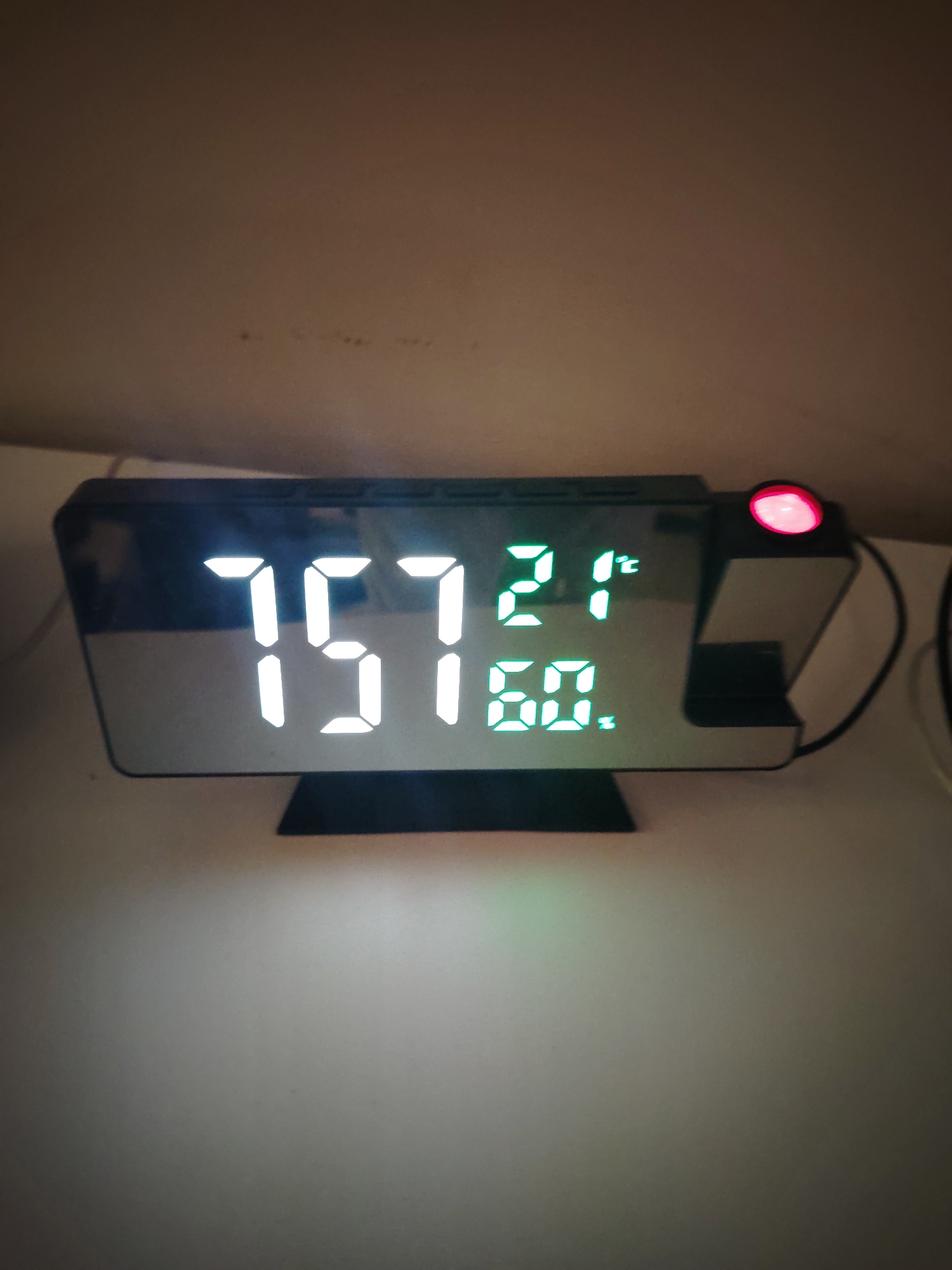 The image captures an indoor scene, located in Huntingdonshire, United Kingdom, featuring a black digital alarm clock with a mirrored face resting on a light-colored surface. The clock's primary display shows '7:57' in prominent white LED digits. To the right of the time, in smaller green LED digits, the temperature '21°C' and humidity '60%' are displayed. A spherical red light, characteristic of a projection mechanism, is illuminated on the clock's upper right side. A black power cable extends from the right side of the device. The background consists of a plain, muted brown wall with minor surface inconsistencies. No individuals are present in the image. The ambient lighting is dim, making the clock's illuminated display the brightest element.