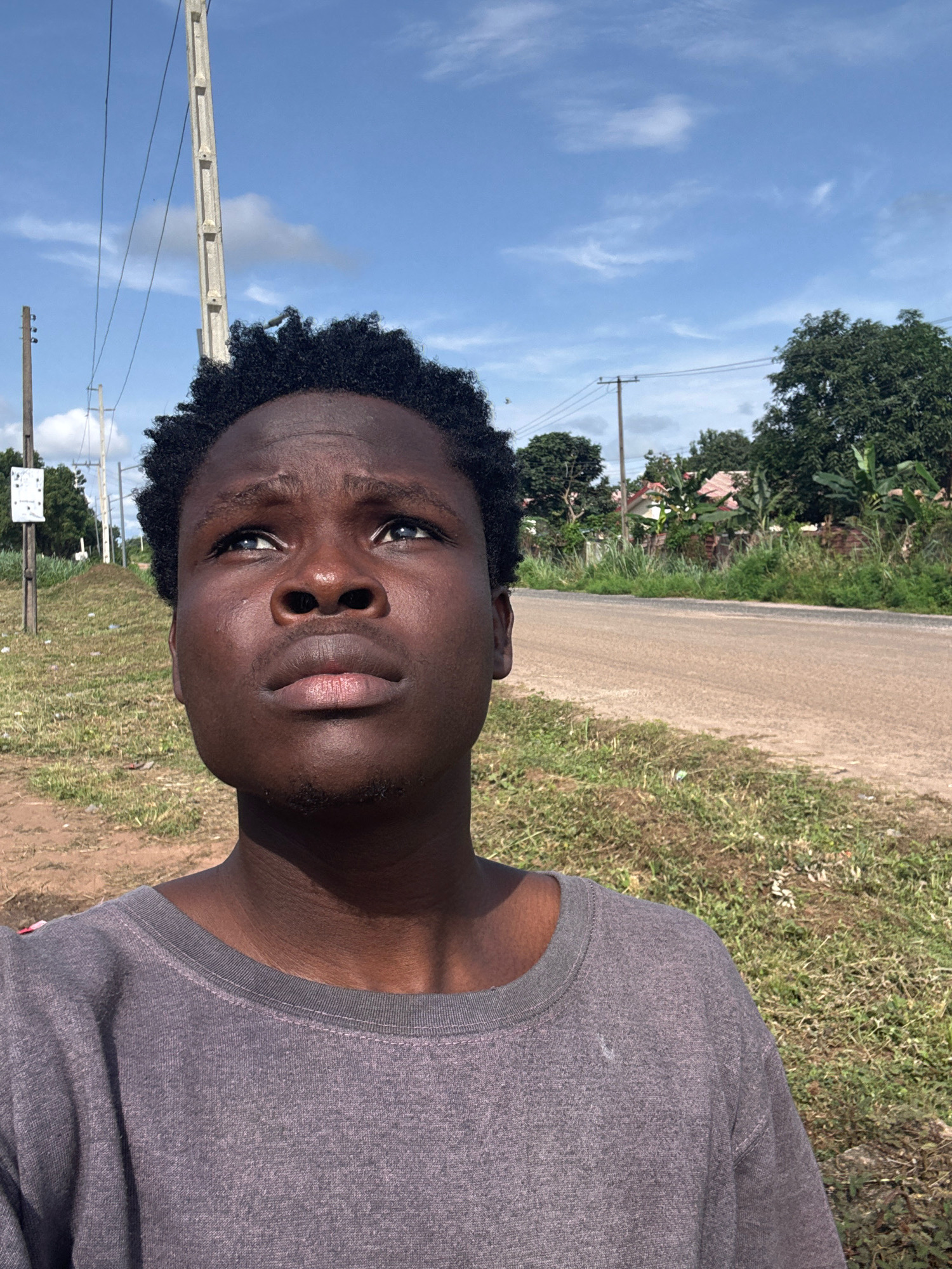 A young man stands outside, looking upwards. He has dark skin and short, curly black hair. He is wearing a grey t-shirt. Behind him is a dusty road, bordered by dry grass and some vegetation. Further in the background, there are trees and houses, suggesting a rural or suburban setting. A telephone pole with wires is visible to the left of the frame. The sky is blue with scattered clouds. The lighting suggests it is daytime. The image appears to be taken in Ihiagwa, Nigeria, as per the context provided.