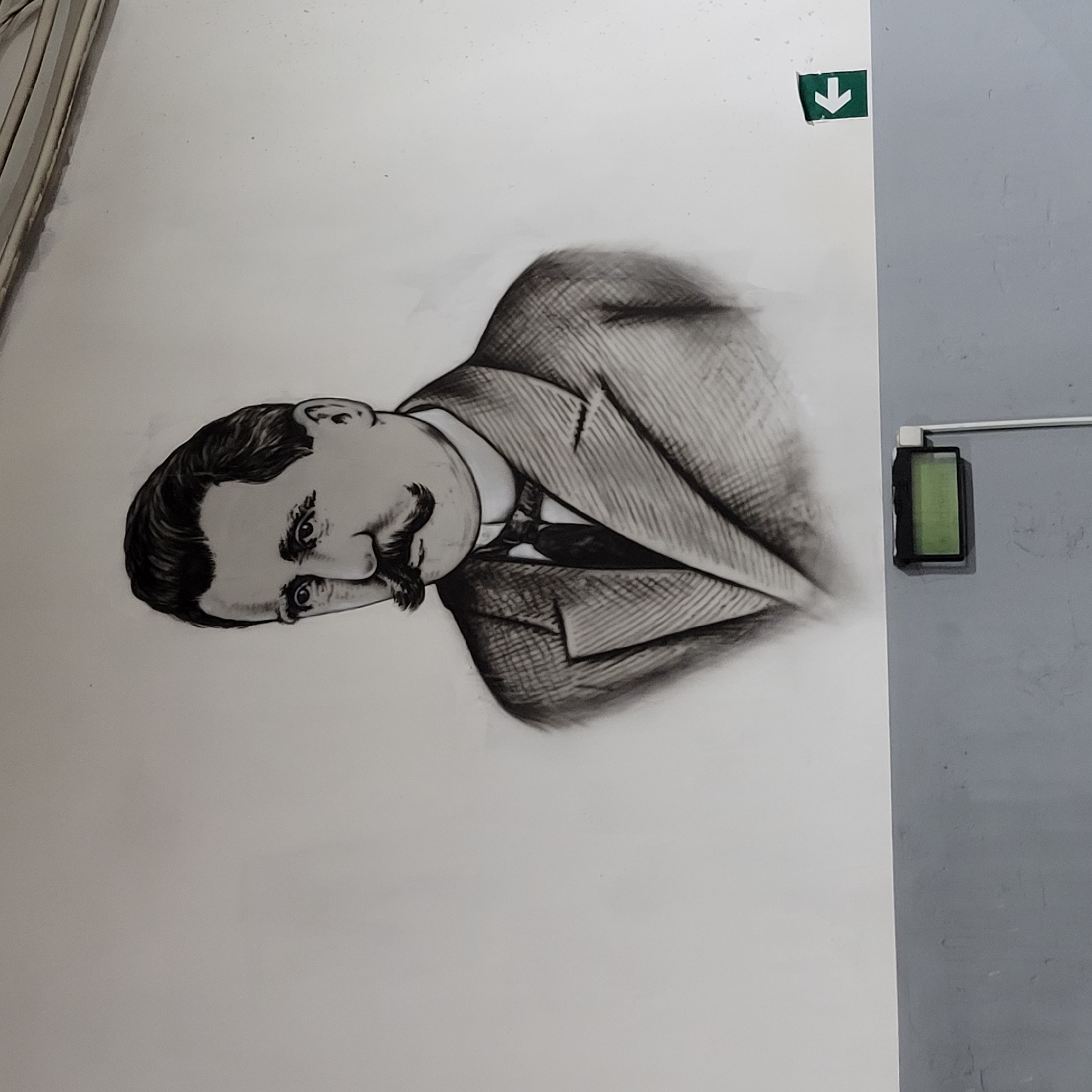 The image presents an indoor scene featuring a wall adorned with a large, black and white illustration of a man. The man, depicted from the chest up, has dark hair, a prominent mustache, and is wearing a suit jacket over a collared shirt and tie. His gaze is directed downwards and slightly to the left, conveying a neutral or contemplative expression.
The illustration is situated on a light-colored wall, likely white. In the upper right section of this wall, a small, square green sticker with a white downward-pointing arrow is affixed. To the right of the illustration, the wall transitions to a grey color. On this grey wall, a black rectangular device with a green illuminated screen is mounted. A slender white bar extends upwards from this device. The arrangement suggests an interior setting, possibly a hallway or public space. No specific activity is occurring; the image captures a static display on the wall.