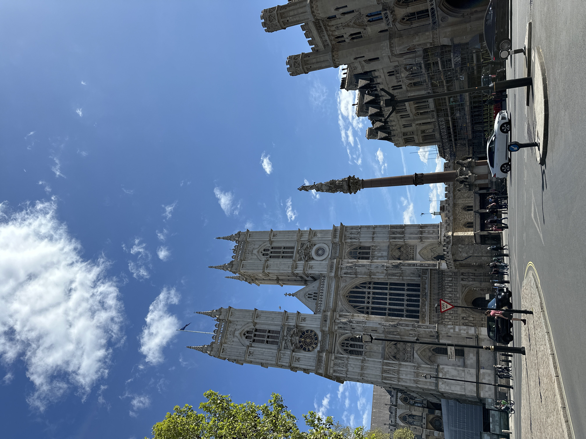 The image shows a view of Westminster Abbey in London, United Kingdom. The sky is blue with scattered clouds. The Gothic architecture of the Abbey is prominent. A monument or column is visible near the Abbey. On the right side, there's a street scene with vehicles and pedestrians, alongside part of a building potentially associated with Parliament.