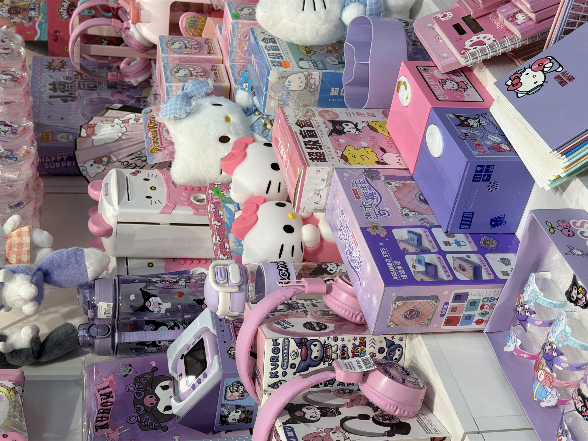 A vibrant, overhead shot displays a dense and colorful collection of Sanrio-themed merchandise, primarily featuring characters like Hello Kitty, Kuromi, and Cinnamoroll. The items are tightly packed on what appears to be a retail shelf or display table, creating a cluttered yet appealing aesthetic.
Dominant objects include numerous plush toys, such as a large white Cinnamoroll with blue gingham details and smaller Hello Kitty heads. A variety of stationery items like notebooks and binders, many featuring Hello Kitty, are stacked. Several "SUPER BLIND BOX" products are visible, showcasing various Sanrio characters including Kuromi, Pompompurin, and My Melody. The boxes also display Chinese text ("超级盲盒").
Multiple pairs of pink and purple headphones are scattered throughout, some still in packaging, with one pink pair priced at "1080 руб." Another pair is labeled "AH-902Y KUROMI SWEET BABY." Purple water bottles featuring Kuromi designs are present, one with a "480 руб." price tag. Other items include a pink Hello Kitty-themed money box, a purple Kuromi-branded electronic device with a screen, and a "DIY ASSEMBLY STES" kit for making accessories. Stacks of small plastic containers with Hello Kitty designs line the left side, and a display of colorful character bracelets is on the far right.
The scene depicts a retail environment, likely a store specializing in character goods. The presence of prices in rubles ("руб.") indicates a location in Russia, aligning with the context of Izhevsk. The artificial lighting suggests an indoor setting. Visible text includes "HELLO KITTY," "KUROMI," "SUPER BLIND BOX," "ASSEMBLY STES," "HAPPY SURPRISE," "LOVED," and "Princess Kitty." The overall impression is one of a cheerful and appealing collection of character merchandise, designed to attract fans.
