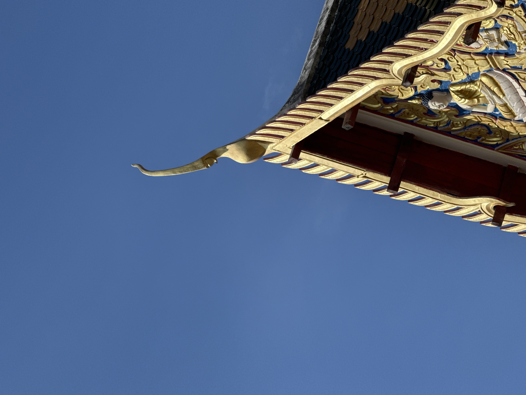 An exterior, low-angle, close-up shot captures the corner of a Thai temple roof against a clear blue sky. The roof is highly ornate, featuring layers of gold and red decoration. The eaves are lined with gold trim. Above this, there are intricate gold carvings of figures against a blue background. At the far left of the image, a curved golden architectural element extends outwards. The photo was taken in Baan Taling Ngam, Thailand.