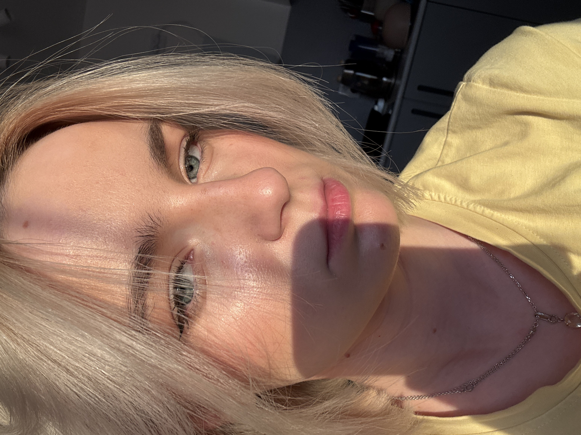 A close-up, angled shot depicts a blonde woman. The woman has fair skin, blue eyes, and light eyebrows. She wears a yellow shirt and a silver necklace. The photograph is taken outdoors, with strong sunlight casting shadows across the woman's face. The background is blurred and indistinct, showing what appears to be an interior space with dark shelving units. The location in the city of Sinyavino, Russia is not discernible from the image.