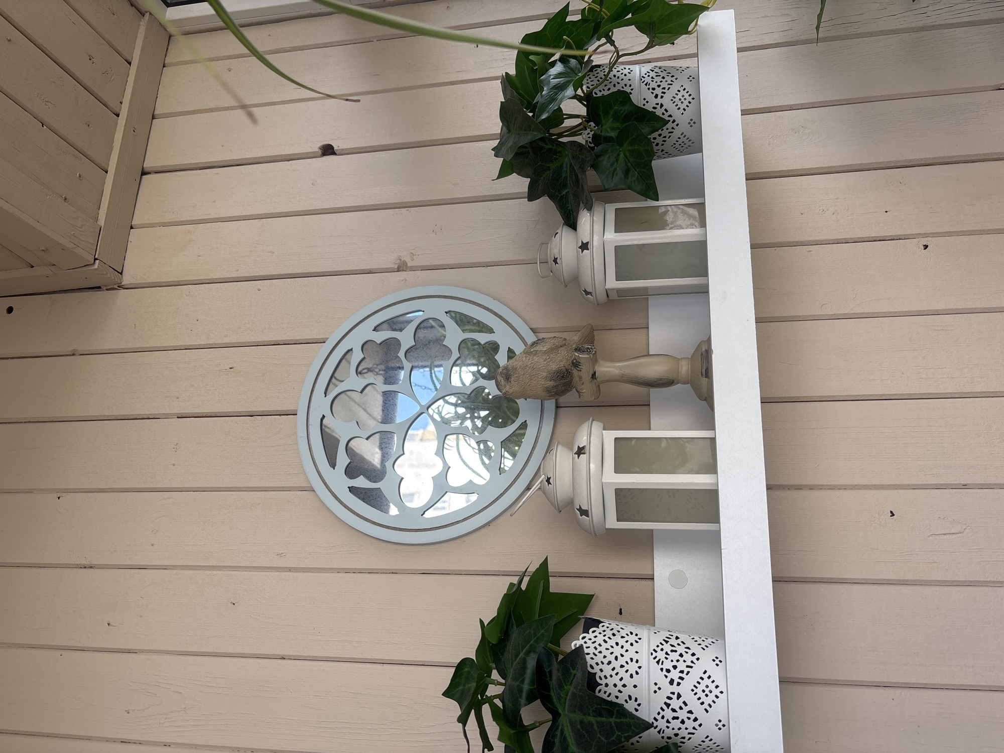 This media file shows a decorative arrangement on a light beige wooden wall, likely an outdoor setting. The wall is made of horizontal planks. Mounted on the wall are several white decorative items, including a round mirror with an ornate cutout frame and several lanterns. Two of the lanterns are visible, stacked vertically on a white shelf. The top lantern has small star cutouts, while the bottom one has a more intricate lattice pattern. A potted plant with green ivy is placed in the top lantern, with its leaves trailing down. Another decorative white pot with a similar lattice pattern is visible at the bottom of the shelf, also containing ivy.
A distinct decorative element is a light blue, circular mirror with a detailed, gothic-style cutout frame that resembles a stained-glass window. A small, decorative object that appears to be a carved animal head is attached to the top of the mirror. The arrangement suggests a focus on decorative outdoor accents. The lighting appears natural, suggesting daytime, and there are no indications of specific weather conditions. The overall impression is one of a charming and aesthetically pleasing display in an outdoor living space.