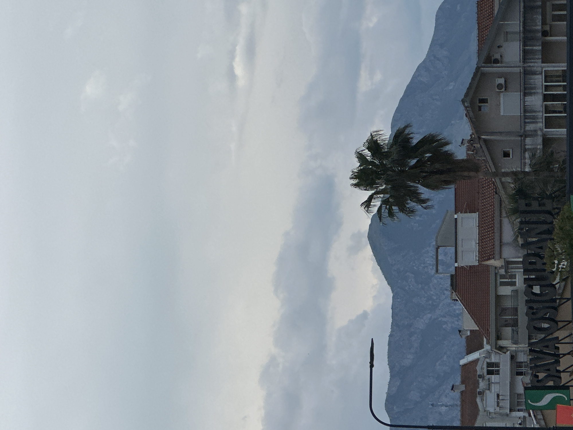 The image depicts a partially cloudy sky above a cityscape with mountains in the background. In the foreground, there is a palm tree and several buildings with red-tiled roofs. A street light is visible on the bottom right. A sign with the word "SAVOŠIĆI-GRAND" is partially visible on the right side. The location is Donji Pobori, Montenegro.