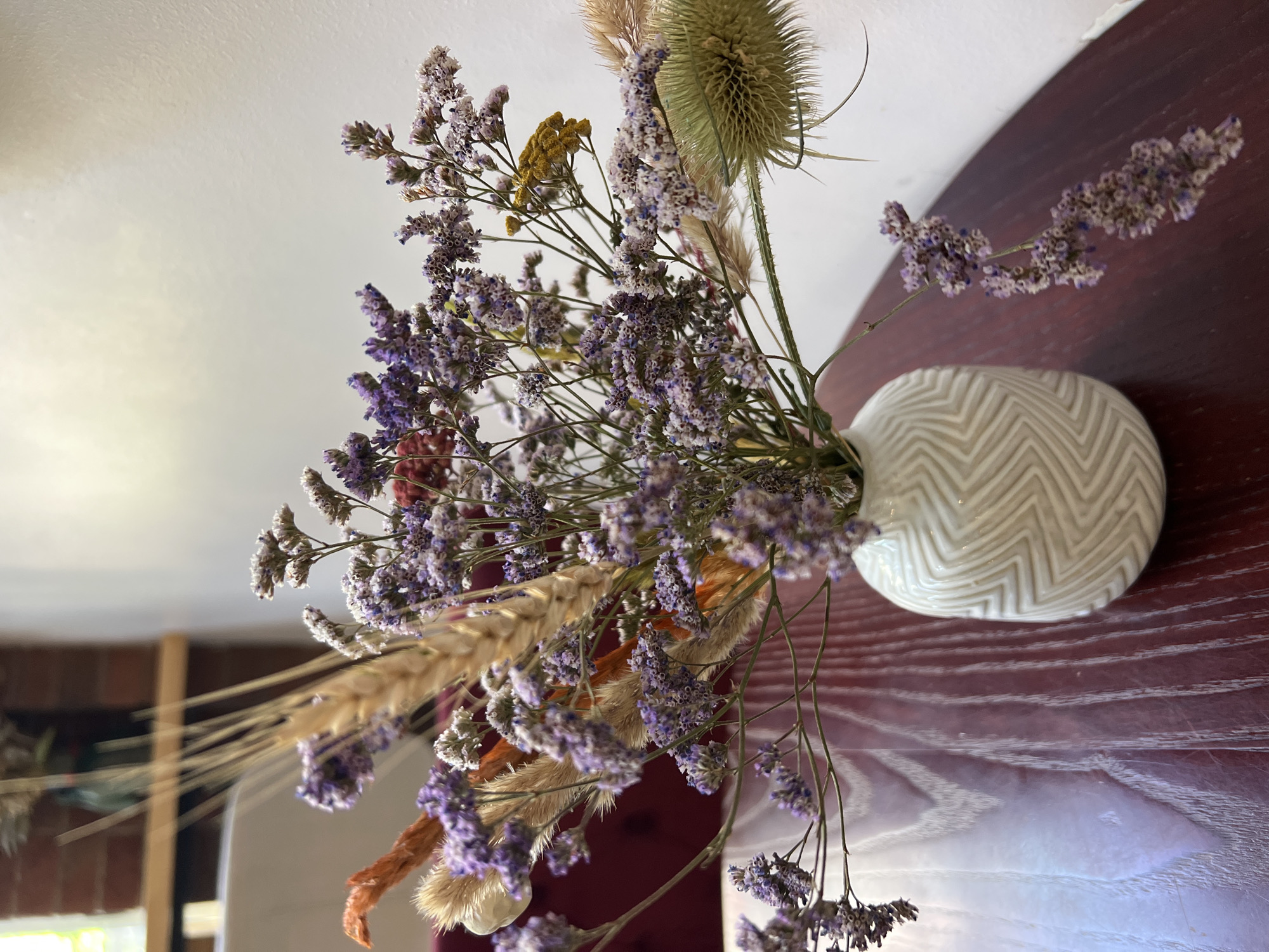 A close-up, eye-level shot shows a dried floral arrangement in a white ceramic vase on a polished dark wood table. The vase, which is cylindrical with a textured, zigzag pattern, sits to the right of the frame. The dried flowers are a mix of purple statice, wheat stalks, and other dried grasses and seed pods, with hints of orange and yellow. The background is blurred, with a white wall on the right and a dark, out-of-focus area on the left that appears to be a room with brick and a window. The lighting is soft and natural, casting gentle shadows. The image is crisp and detailed, focusing on the textures and colors of the dried flowers and the vase. There are no people or animals present in the scene. The location is identified as Fontanka, Ukraine.