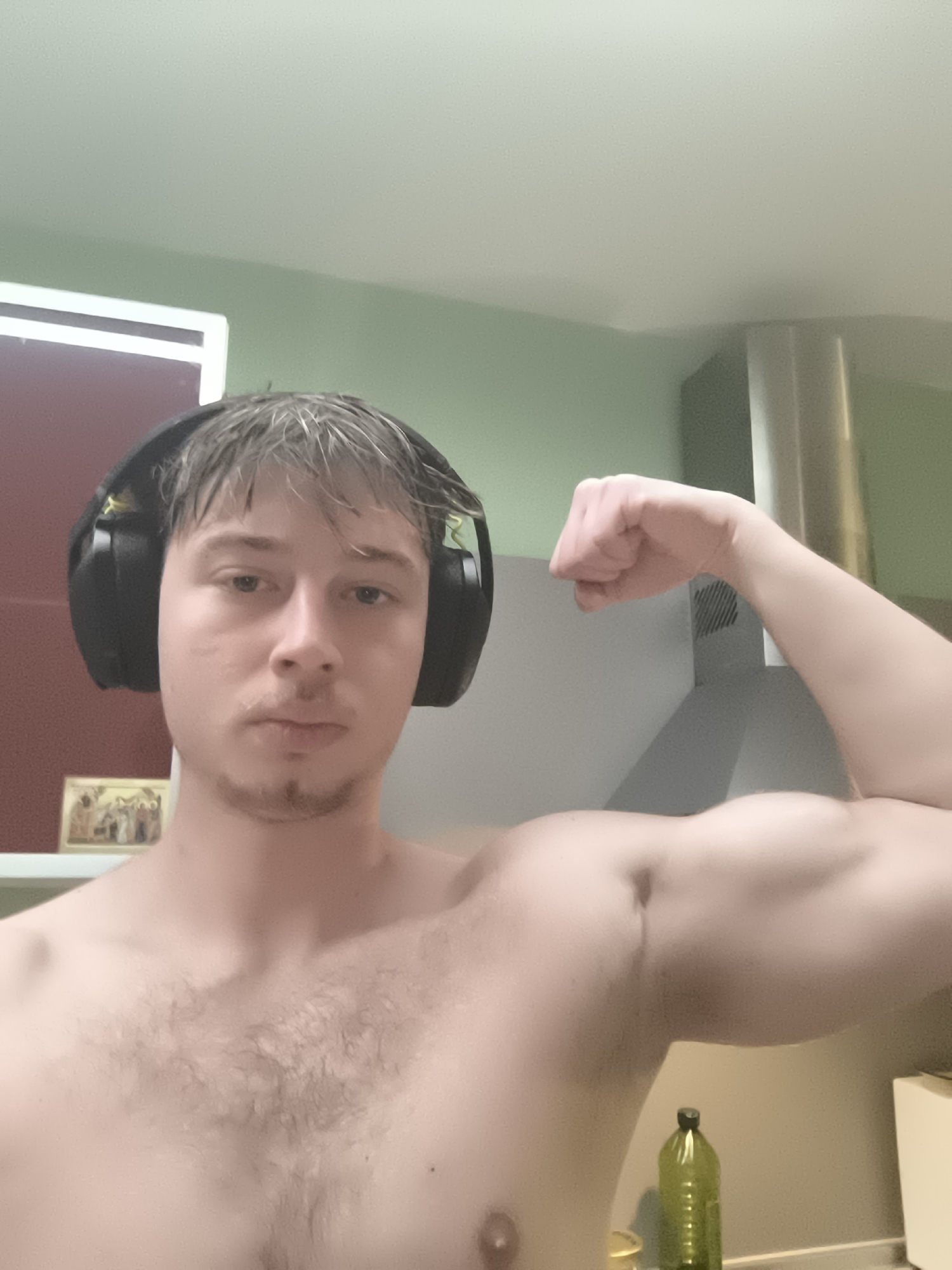 A shirtless young man with short, blonde hair and stubble poses for a selfie while wearing headphones. He is flexing his right bicep. The background includes a kitchen area with an olive green wall, a stainless steel chimney hood, and a red cabinet. A green plastic bottle is visible on the counter. The image does not provide sufficient information to determine a specific location in Sofia, Bulgaria.