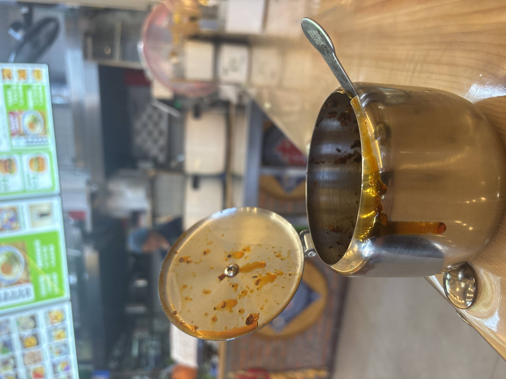 A close-up shot focuses on a metal condiment holder with a spoon resting inside. The holder is partially filled with a reddish-orange sauce, and some sauce has dripped down the side. The lid of the holder is open, revealing residue of the same sauce. In the background, the scene is blurred, showing what appears to be a food service establishment. A blurred figure in a blue cap is visible behind a counter, and menu boards displaying food items are mounted on the wall. The overall setting suggests a casual dining or fast-food environment in Suzhou Industrial Park, China, where customers might access condiments for their meals.