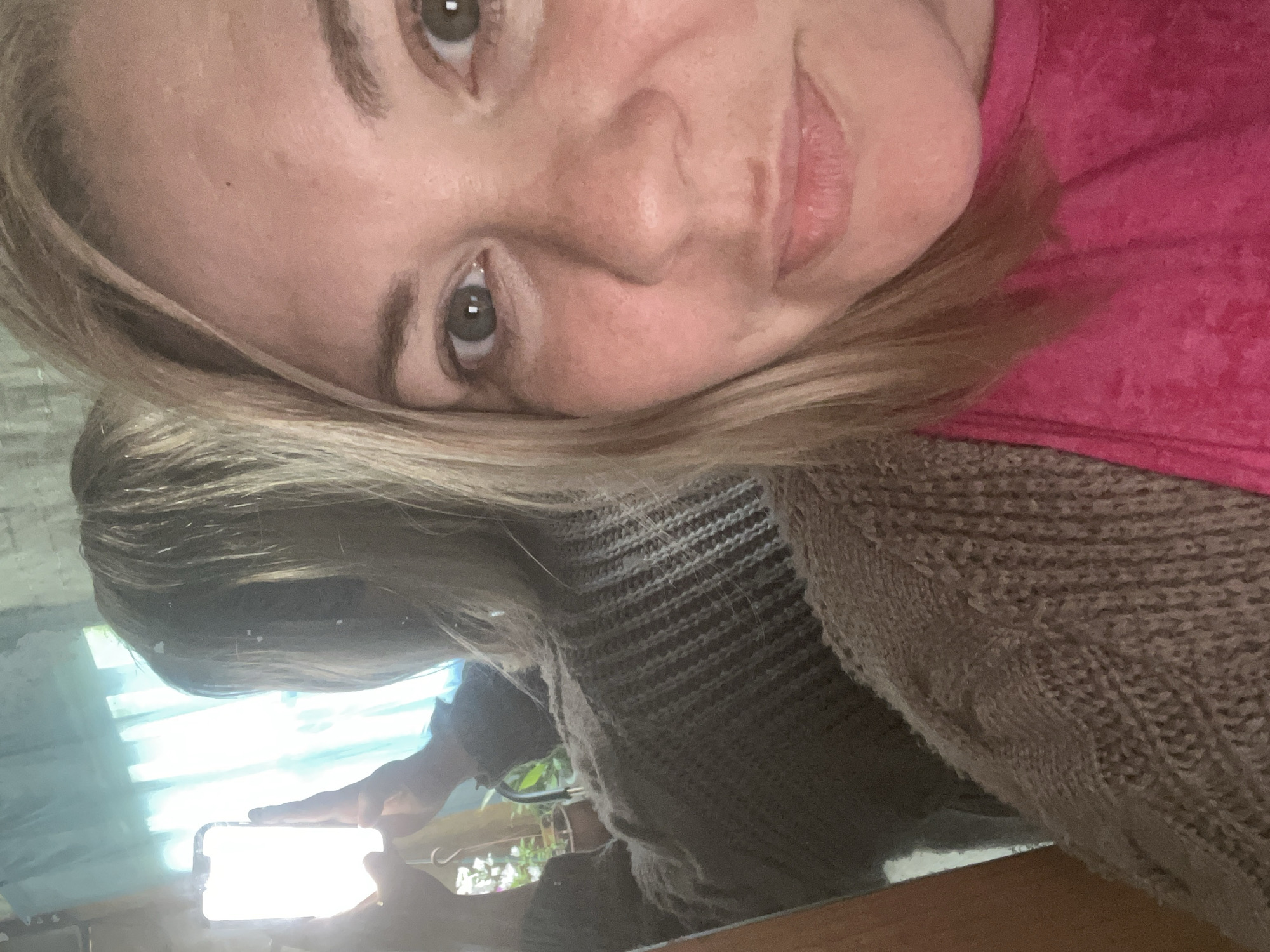 A person is taking a selfie in a mirror. The person has blonde hair, brown eyes, and is wearing a pink top and a brown sweater. The reflection shows a phone being held up, with a bright screen. The background reflection shows a window with light coming through, and a glimpse of plants. There is no information to suggest this image was taken in Vesyoloye, Russia.