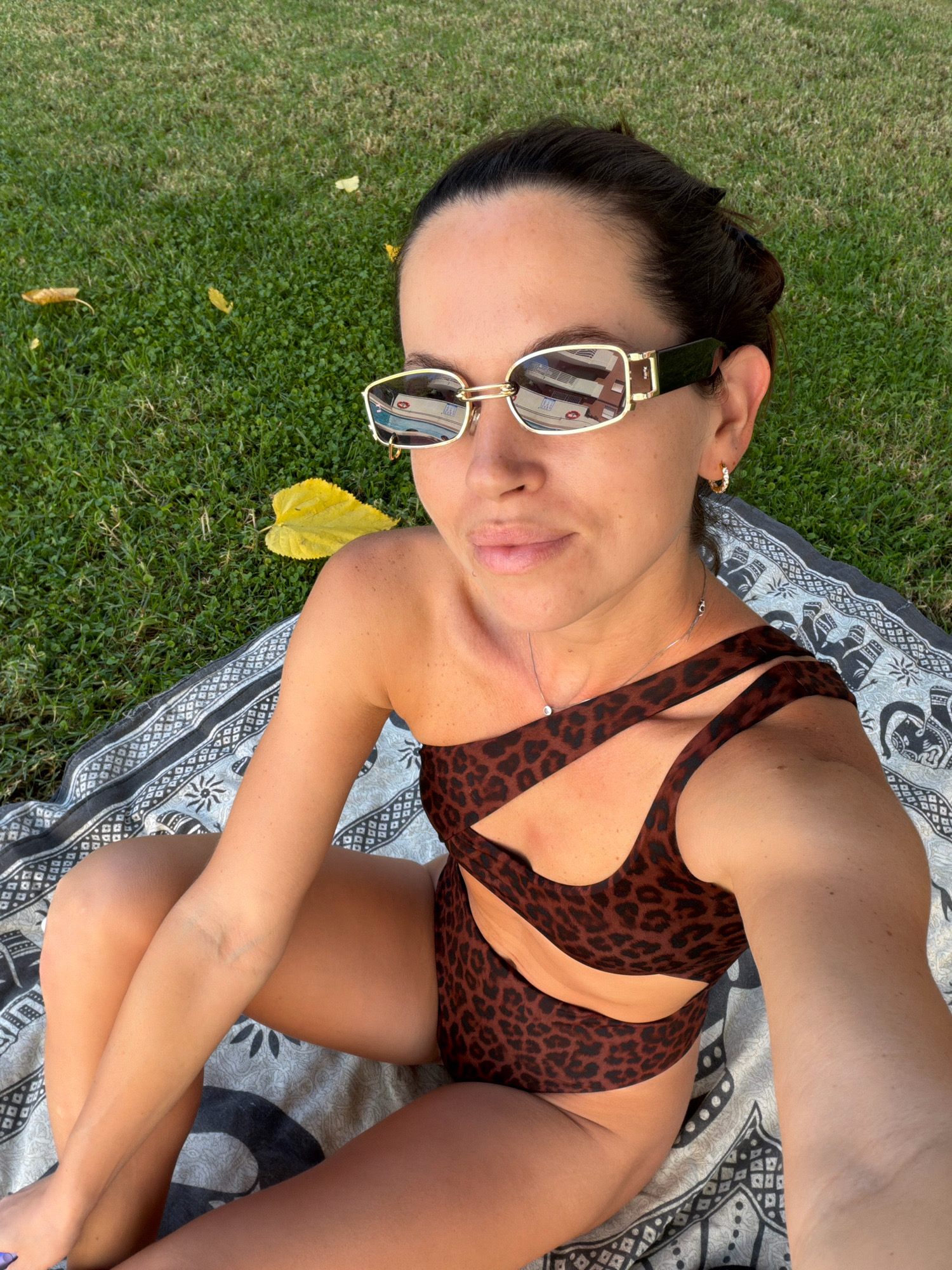 A woman is taking a selfie while sitting on a blanket spread on green grass. She is wearing a leopard-print, one-shoulder swimsuit with cutouts and gold-rimmed sunglasses. Her hair is dark and pulled back. She has a delicate necklace with a small pendant and hoop earrings. In the reflection of her sunglasses, a building and a boat are visible. A large yellow leaf is on the grass near her. The scene appears to be outdoors, likely in a park or backyard, on a sunny day. The time of day is likely afternoon, given the bright sunlight and shadows.