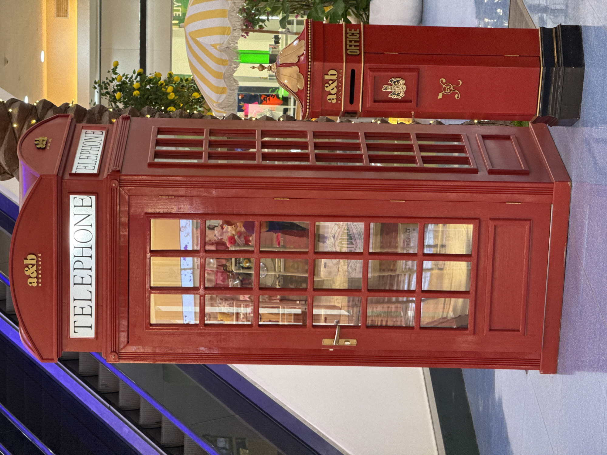 The image depicts a vibrant red, replica British telephone booth positioned next to a matching red, pillar-style mailbox. The telephone booth has a classic design with multi-paned glass windows and a golden door handle. Above the windows, "TELEPHONE" is written in white lettering on a dark rectangular background. A smaller sign with "a&b BOOKCAFÉ" is visible at the top of the booth. The mailbox is adorned with a golden crest and the word "OFFICE". The scene appears to be set within an indoor shopping mall, indicated by the presence of an escalator with purple lighting in the background, potted plants with yellow flowers, and the reflective, blue-toned floor. The lighting is bright and suggests an artificial environment. There are no people clearly visible in the image, though reflections in the telephone booth windows suggest activity within the store it represents. The overall impression is one of a themed decorative installation, possibly part of a shop or a promotional display. The location is specified as Porlamar, Venezuela, suggesting this is a commercial or public space within that city.