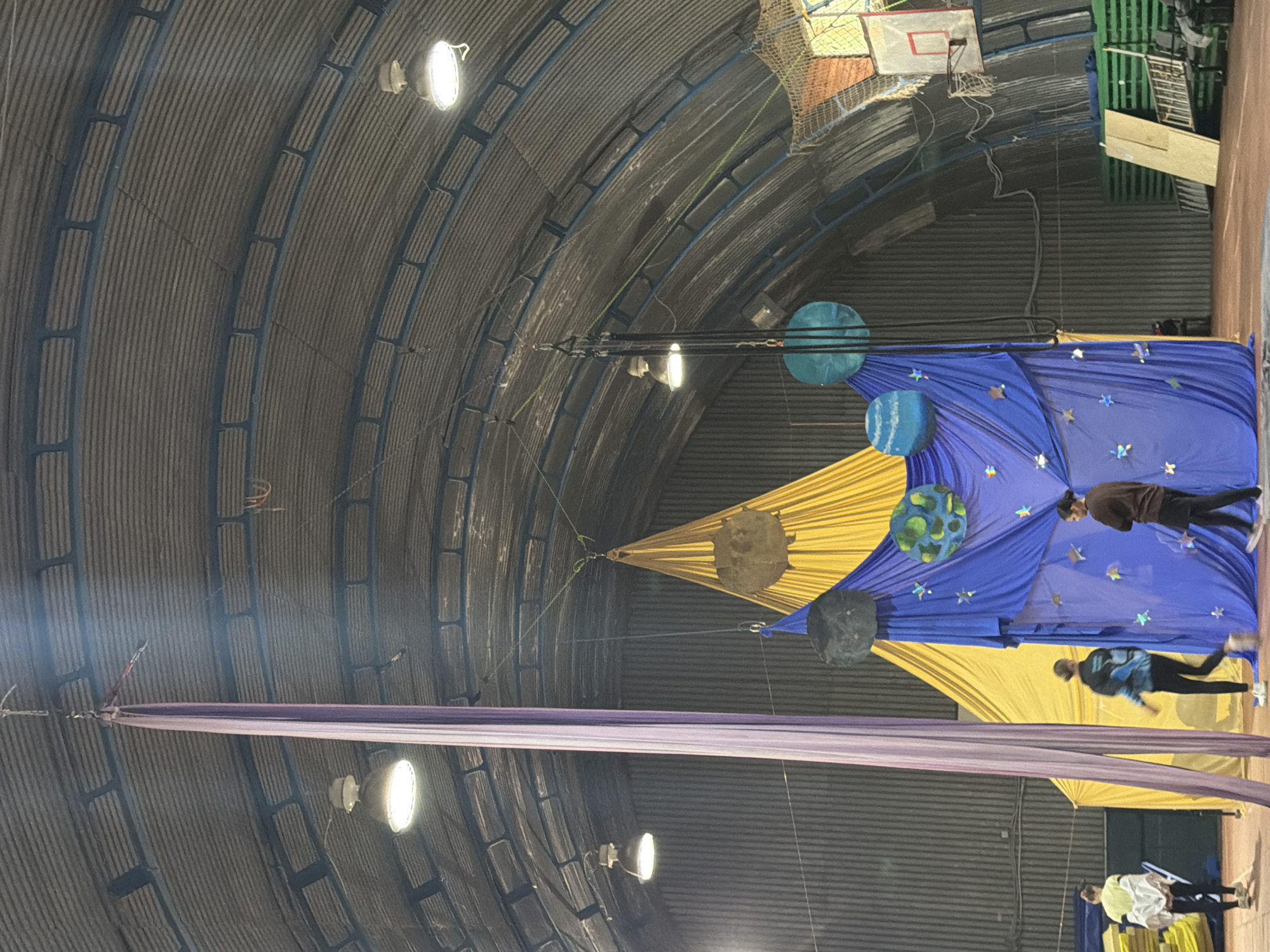 The image depicts an indoor circus or performance space with a high, corrugated metal roof. A long, purple aerial silk hangs from the ceiling on the left side of the frame, alongside a yellow and blue fabric backdrop decorated with stars and planet-like spheres. Two individuals are visible in the foreground, facing away from the camera and looking towards the backdrop. One person, wearing a dark jacket and pants, stands to the right, while another, wearing a blue and black jacket and black pants, is positioned to the left and slightly ahead. A third person in a white jacket and black leggings is partially visible on the far left, walking away from the backdrop. In the background, a basketball hoop is mounted on the wall. The floor appears to be a reddish-brown surface, possibly a sports mat. The overall impression is of a practice or setup for an aerial performance, potentially with a celestial theme.