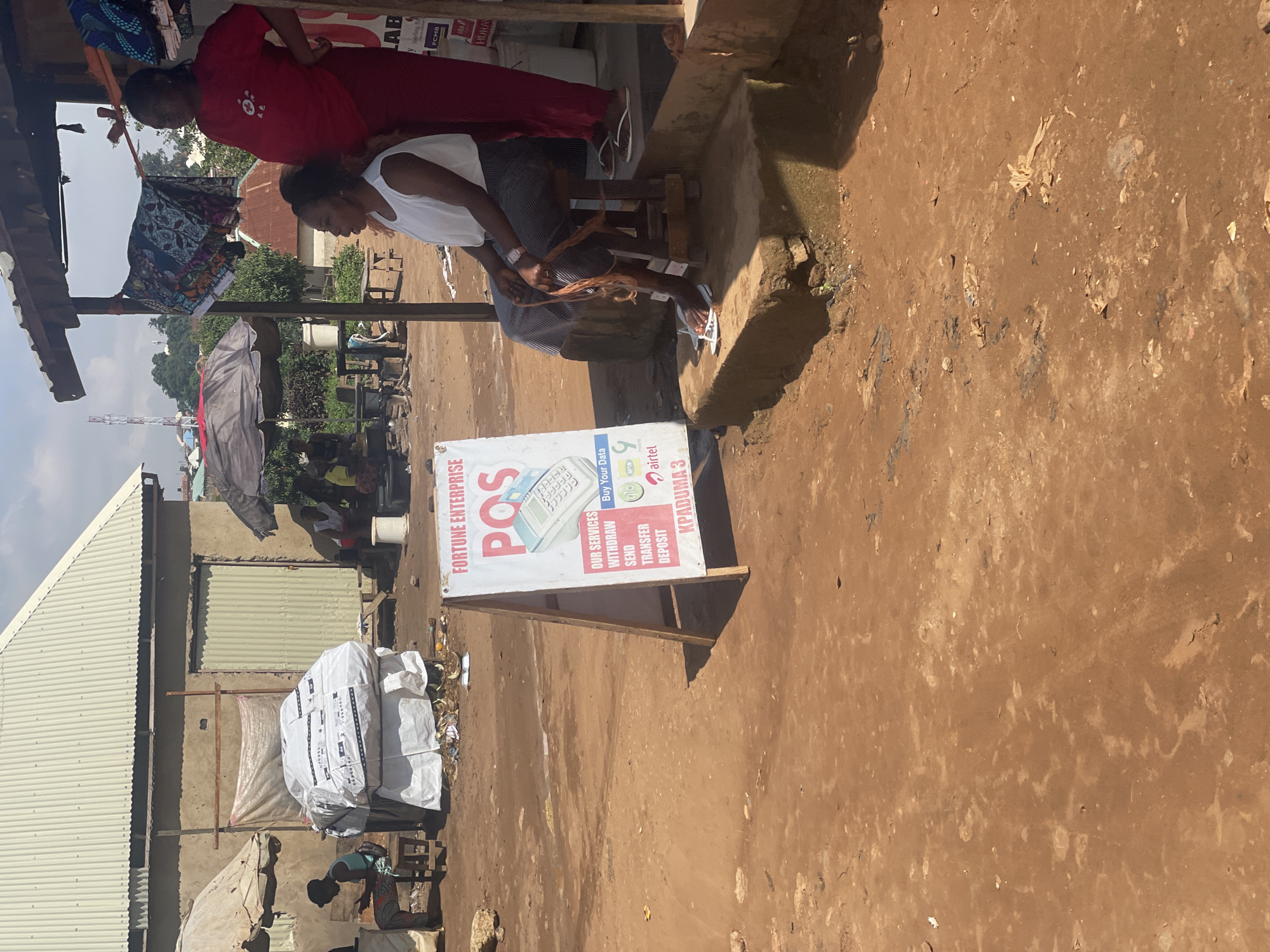In Kurunduma, Nigeria, a dusty, unpaved outdoor scene depicts a commercial area with a focus on financial services. A white A-frame sign for "FORTUNE ENTERPRISE POS" stands prominently, detailing services like withdrawal, send, transfer, and deposit, and advertising "Kpaduma 3." To the right of the sign, two women are visible near an open-air shop. One woman, wearing a red top and dark skirt, stands looking to her left. The other woman, in a white top and striped skirt, sits on a wooden stool, appearing to be engaged in some activity with her hands, possibly with threads or material. In the background, various structures suggest a market or roadside business area, including corrugated metal roofs and awnings. A large, white-wrapped item, possibly goods or packaging, is stacked near a building on the left. The ground is covered in reddish-brown dirt and scattered debris.