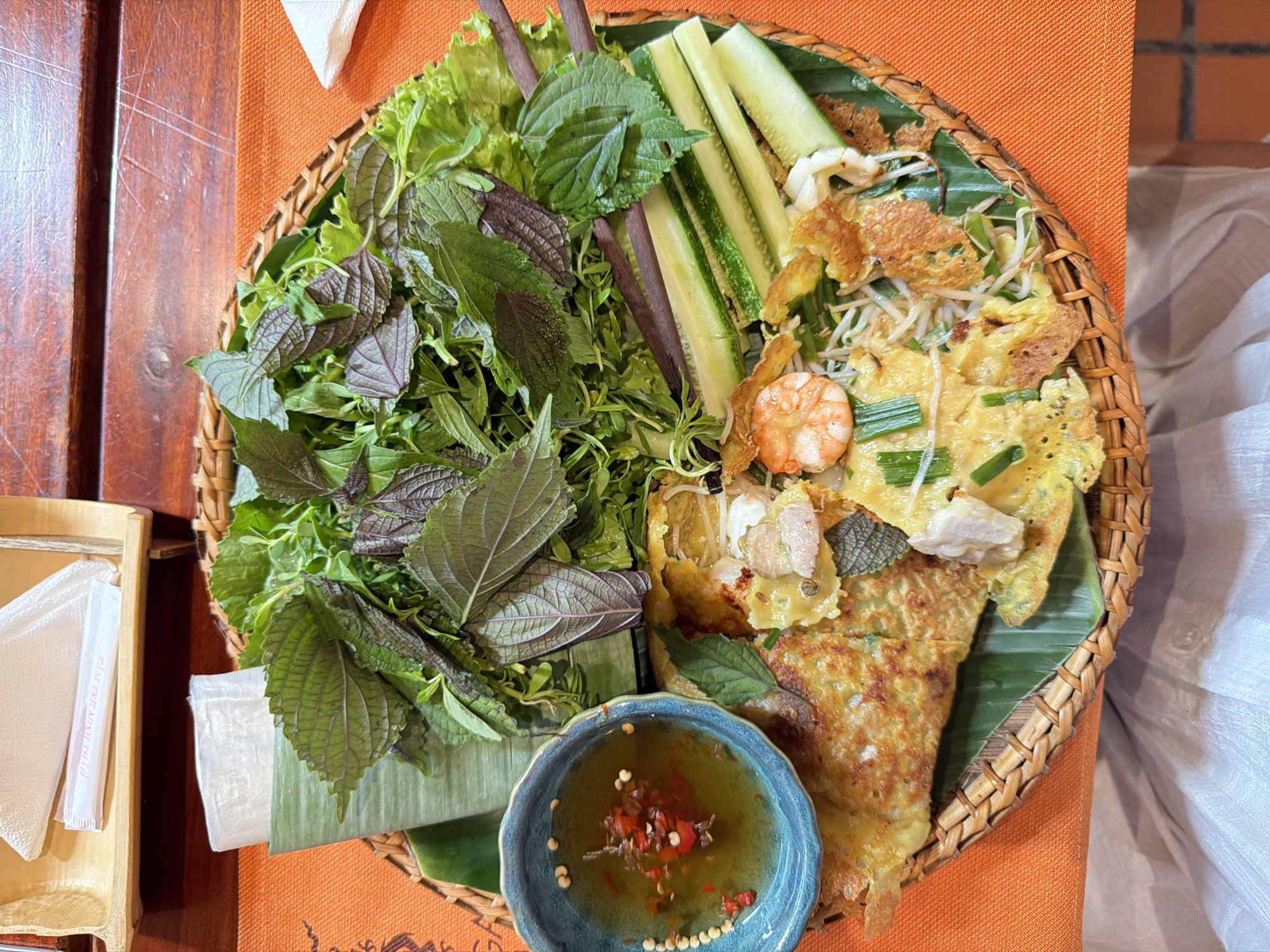 The image shows a close-up, top-down view of a Vietnamese dish, likely Banh Xeo, served on a woven tray with an orange placemat. The dish is a savory pancake filled with shrimp, bean sprouts, and possibly pork, topped with green onions and herbs. It is accompanied by a small blue bowl of dipping sauce, sliced cucumbers, and a generous portion of fresh herbs, including lettuce and what appears to be perilla leaves. In the background, a wooden tray holds a stack of rice paper wrappers and a packet of chopsticks. The setting suggests a restaurant environment in Nha Trang Ward, Vietnam, indicated by the food presentation and surrounding elements. There are no people or discernible actions visible in the image.