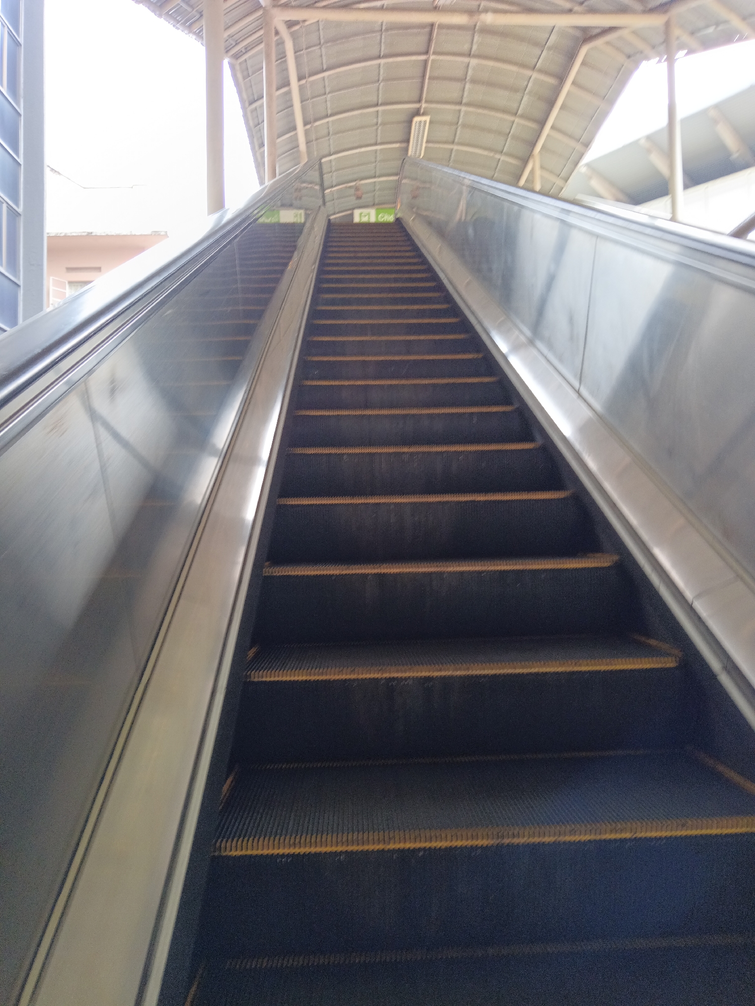 The image shows an upward-facing perspective of a wide escalator in what appears to be a public transportation hub, likely a train station or airport, in Kuala Lumpur, Malaysia. The escalator has dark treads with yellow safety lines. The handrails are metallic and reflective. Above the escalator, a curved, corrugated metal roof structure is visible, supported by industrial-style beams and columns. The sky is bright and washed out, indicating it might be daytime. To the left, a section of a building with blue-tinted windows and a beige facade is partially visible, along with some architectural elements of the station's main structure. There are no people visible in the frame, and the scene is static.