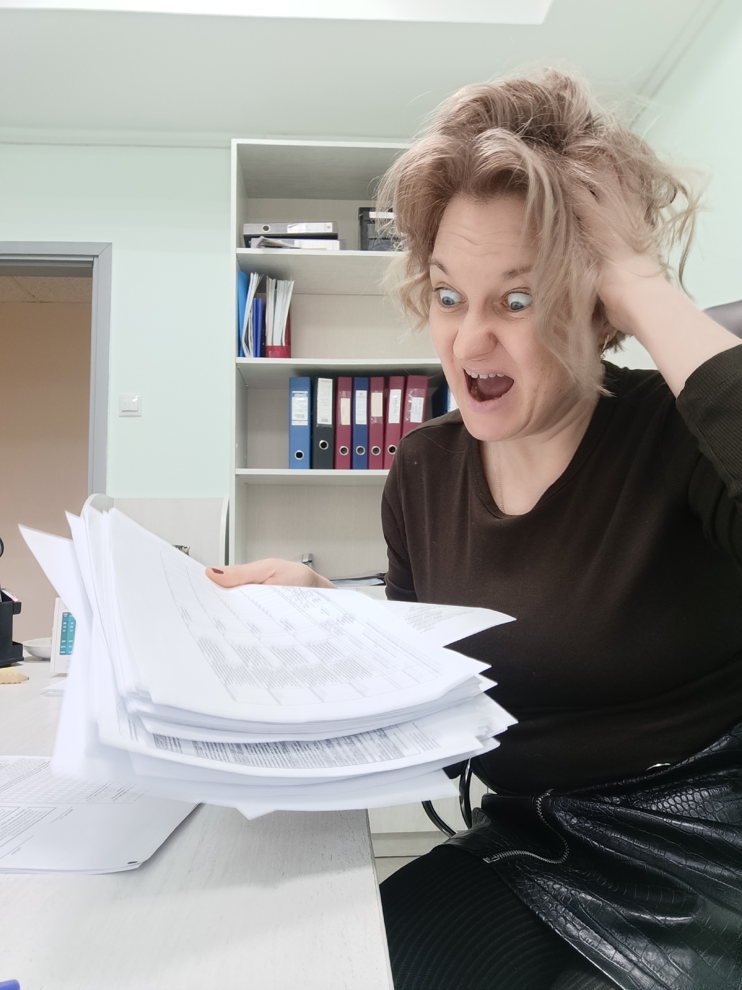 A woman with blonde, disheveled hair sits at a desk in an office setting, appearing distressed while looking at a stack of papers. Her right hand is touching her hair, and her mouth is open in a scream or shout. She is wearing a dark brown sweater and a black textured skirt. The desk is white, and there are additional papers on it. Behind her is a white shelving unit filled with books and binders. The location is likely in Марковский, Russia.
