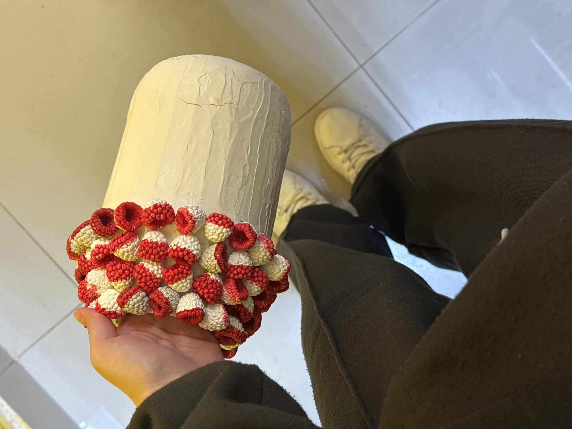 A person is holding a cylinder-shaped object with a textured white surface and a decorative red and white band. The person is wearing dark pants and light-colored sneakers. The setting appears to be indoors, with tiled flooring. The location is Unknown, Unknown.