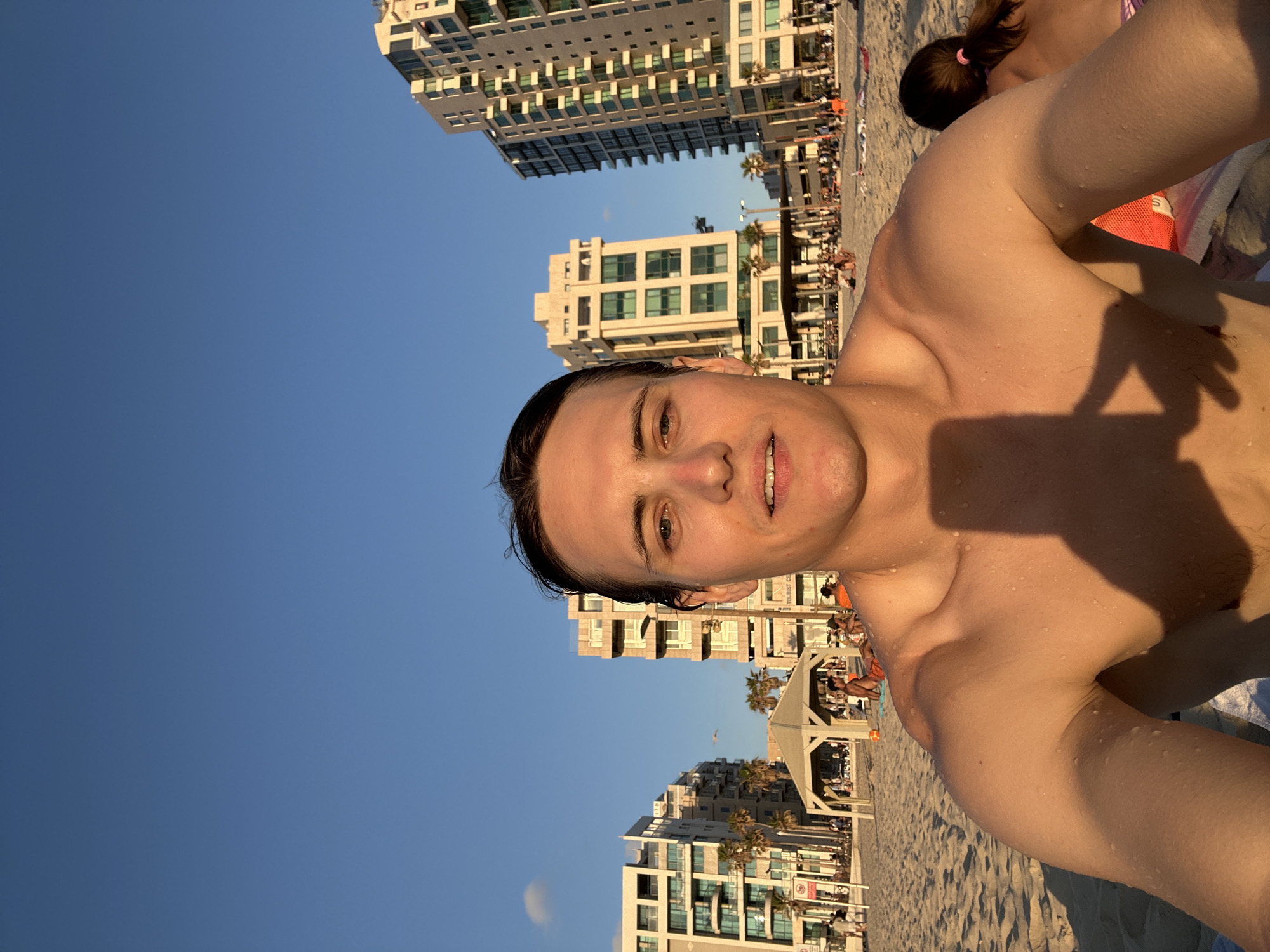 This is a selfie taken on the beach in Tel Aviv, Israel, with the Mediterranean Sea out of frame. The subject is a shirtless man with wet hair, facing the camera. He appears to be smiling. In the background, there are several tall, modern buildings, palm trees, and other beachgoers. The sky is clear and blue, indicating it is daytime. The sand is golden, and there are beach chairs and umbrellas visible. The sun appears to be setting or rising, casting a warm glow on the scene.