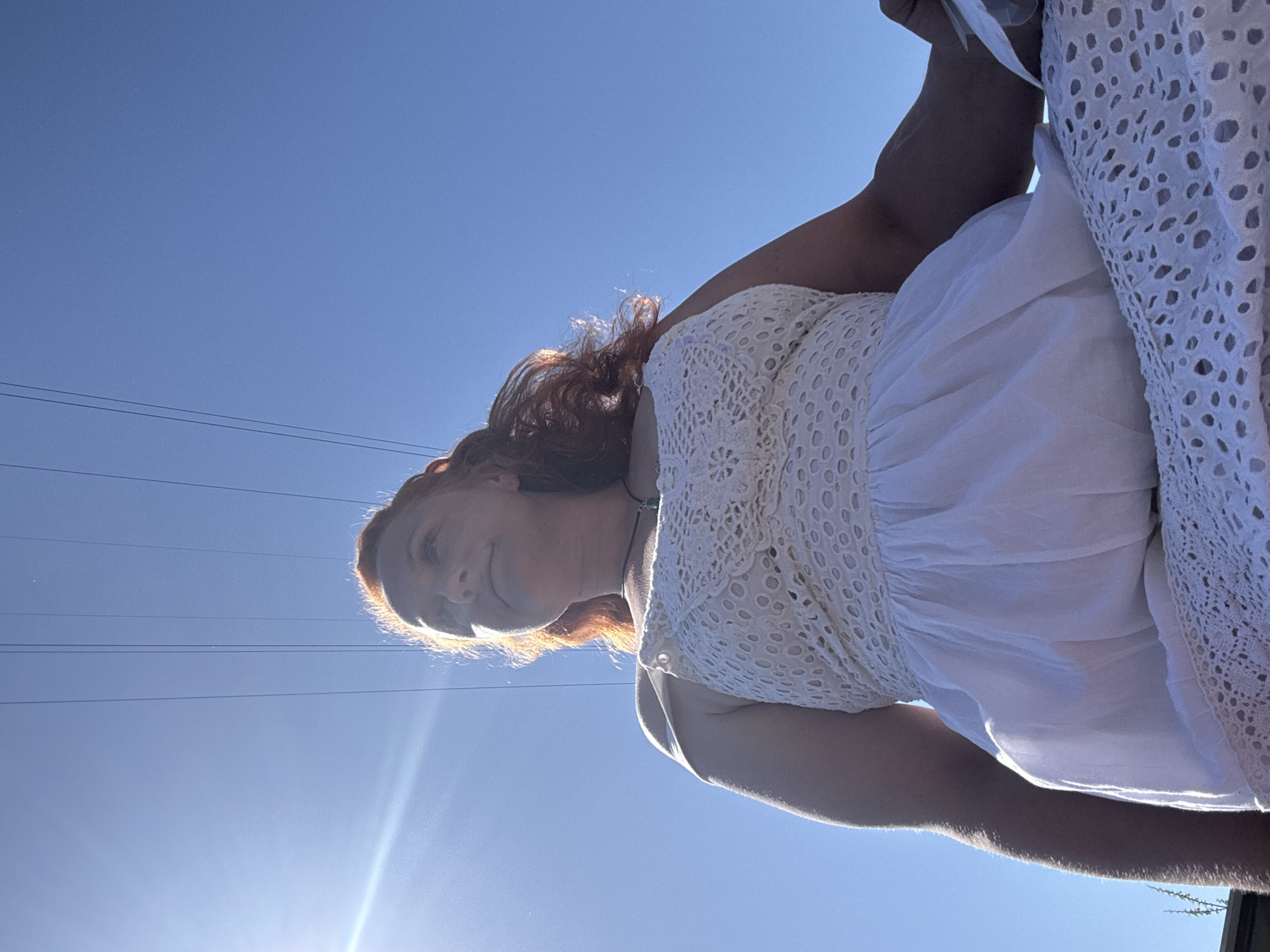 A woman with red hair is wearing a white dress and standing outside on a sunny day in what appears to be an outdoor setting. The sky is bright blue, and power lines are visible in the upper-left corner of the frame. The location in Chaikovsky, Russia, cannot be definitively determined from the visual information provided.