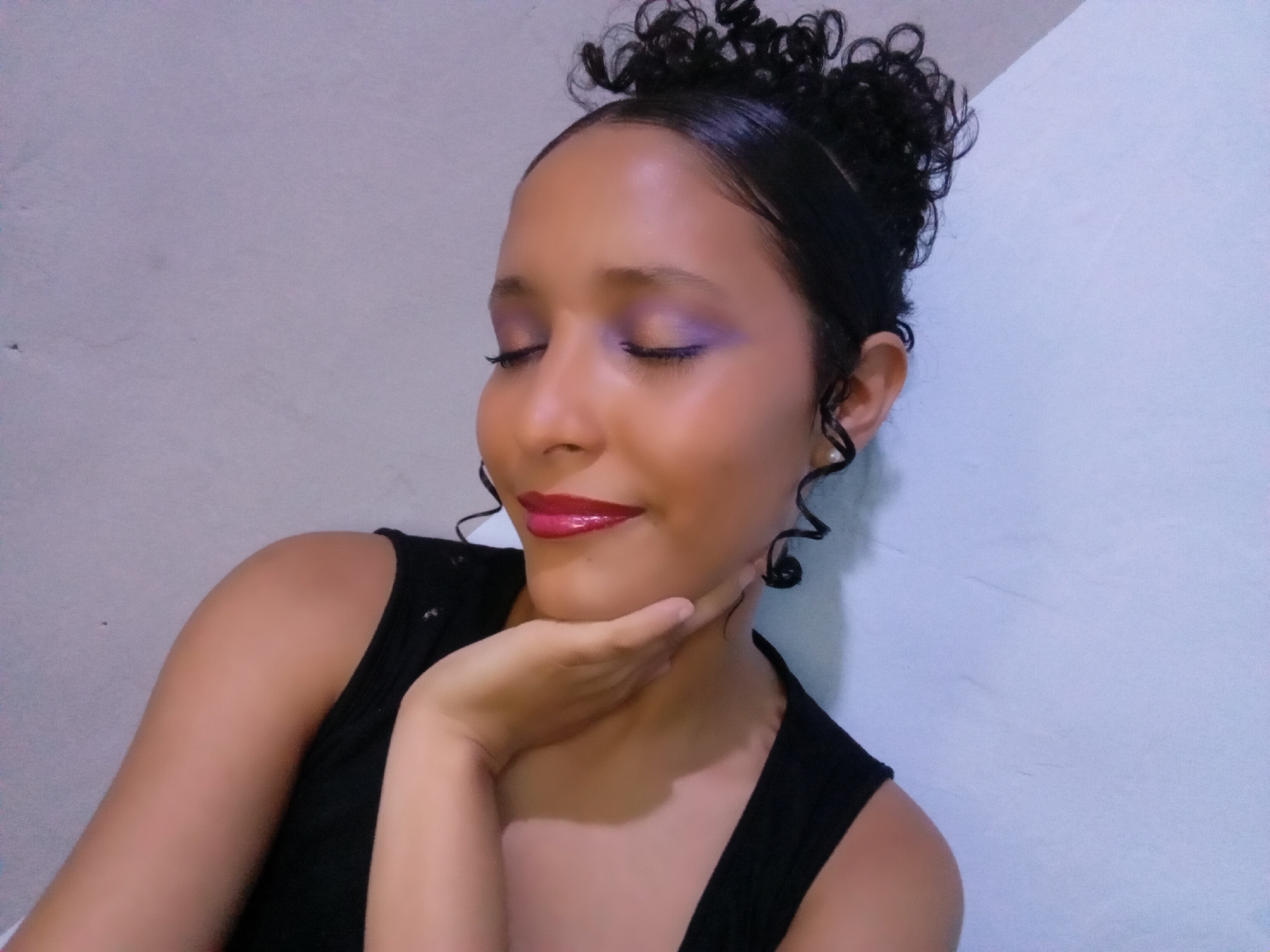 A young woman with dark, curly hair styled in a high bun and artfully arranged tendrils poses in a close-up shot. Her eyes are closed, and she holds her left hand gently cupping her chin, displaying glossy, bright red lipstick and shimmering purple eyeshadow. She has a serene, gentle smile on her face. Her skin appears smooth and radiant under what seems to be soft indoor lighting. She is wearing a simple black sleeveless top. The background consists of a plain, light-colored wall, possibly with a subtle texture or light variations in tone, shifting between light gray and hints of pale blue or lavender. The overall mood is one of tranquility and self-expression. The image was taken in Parroquia Ocumare, Venezuela. No text is visible in the image.