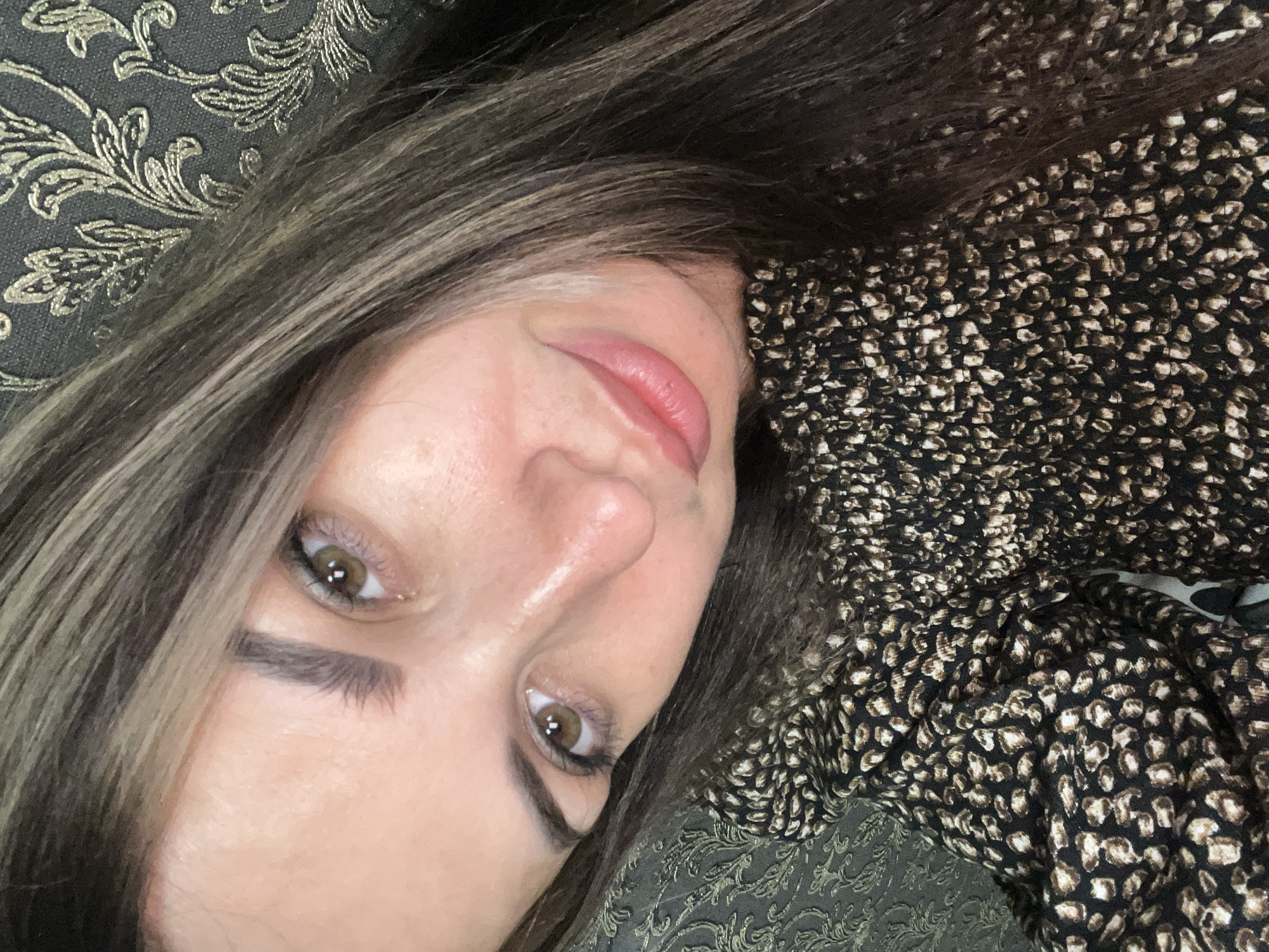 A woman is captured in a close-up, self-taken photograph, her head tilted and resting on something soft. She has long, dark hair with lighter streaks, which falls around her face. Her eyes are brown, and her eyebrows are well-defined. A subtle smile graces her lips, which are painted a soft pink. She is wearing a patterned top with a black base and a repeating leopard-like print of brown and tan shapes. The background appears to be a textured wallpaper with a damask-like pattern in muted tones of gray and gold. The lighting is soft, highlighting the contours of her face and creating a gentle glow. The overall impression is intimate and personal.