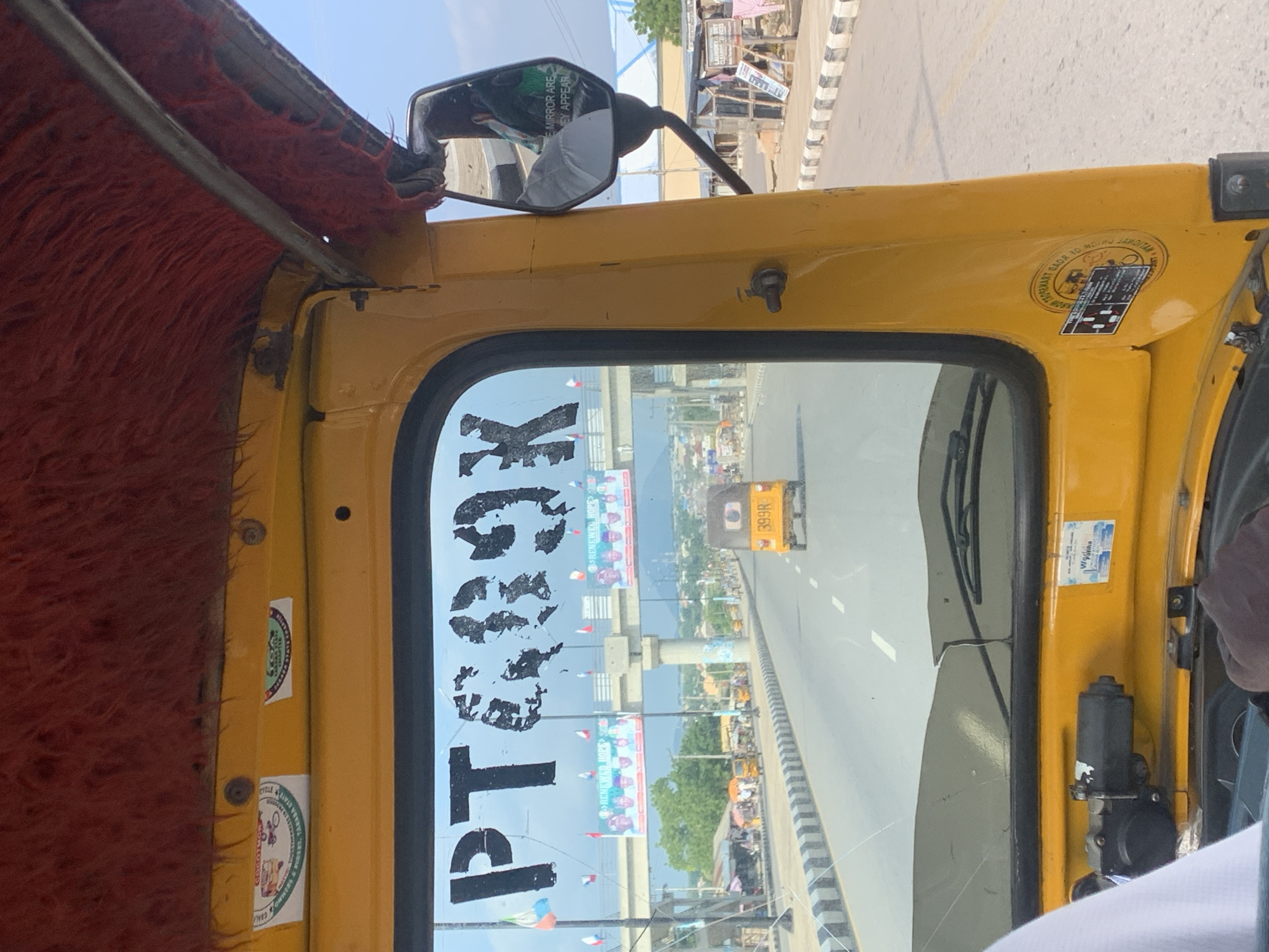 A yellow auto-rickshaw, identified by the license plate "PT 689K," is traveling on a paved road in Jalingo, Nigeria. The auto-rickshaw's interior features a red fuzzy headliner and a side mirror. On the windshield, the license plate is prominently displayed, along with what appears to be stickers. In the foreground, a portion of a white garment is visible, suggesting the perspective of a passenger. The road is flanked by a sidewalk with a black and white striped curb. In the background, other vehicles, including another yellow auto-rickshaw with license plate "399R," are visible. Buildings and a bridge are present along the roadside, with banners displaying political figures and the slogan "RENEWED HOPE" attached to the bridge. The sky is clear and blue. The environment suggests a sunny day in a moderately populated urban or suburban area.