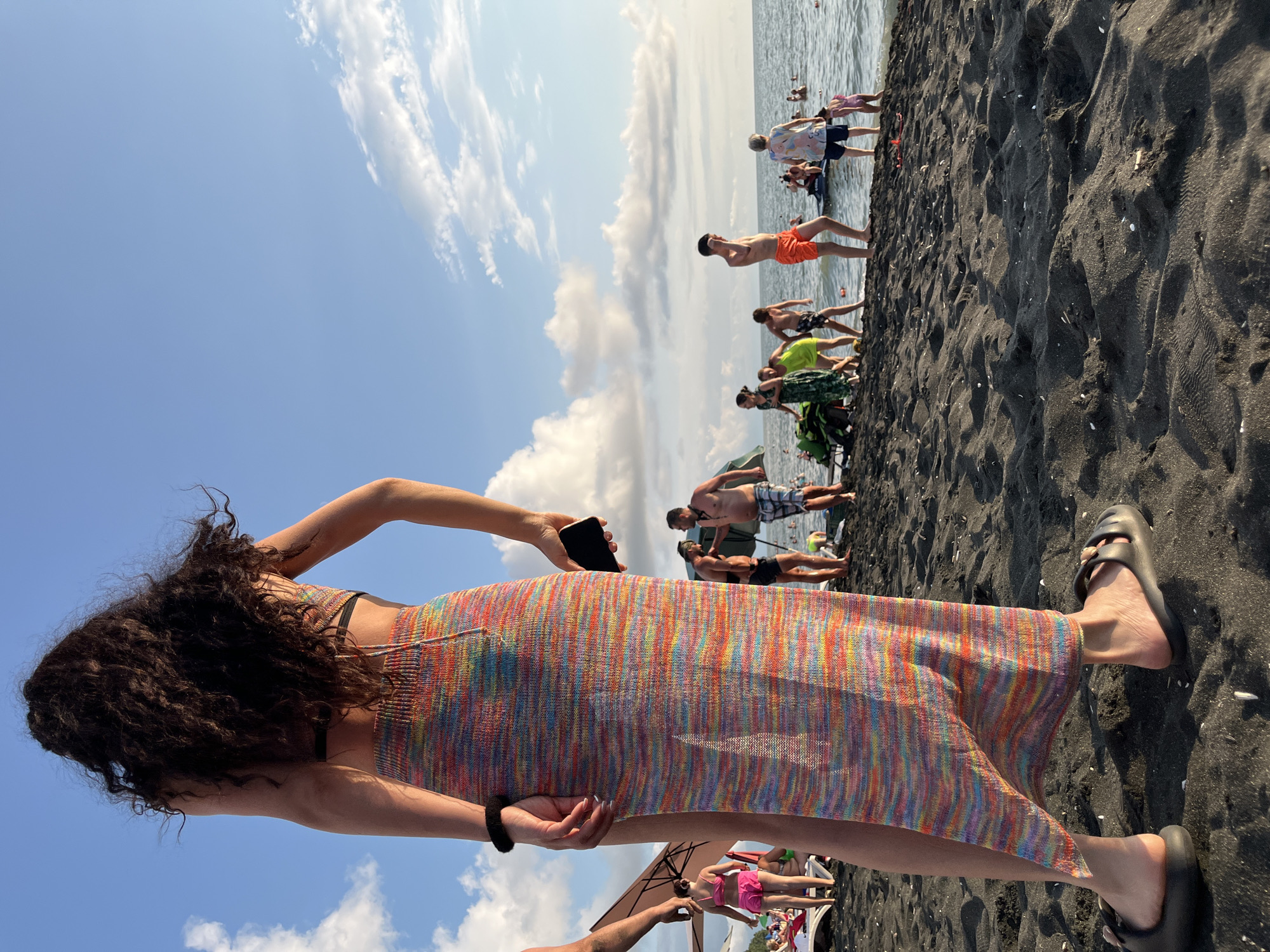 In Ureki, Georgia, a sandy beach is captured from a low angle, focusing on a person from behind wearing a colorful, knitted maxi skirt and dark sandals. The person is holding a black smartphone in their right hand. The background features a clear blue sky with scattered white clouds. The beach is populated with numerous people, some swimming in the sea, others walking along the shore or relaxing. Several beach umbrellas are visible. The sand is dark, typical of Ureki's magnetic sand beaches.