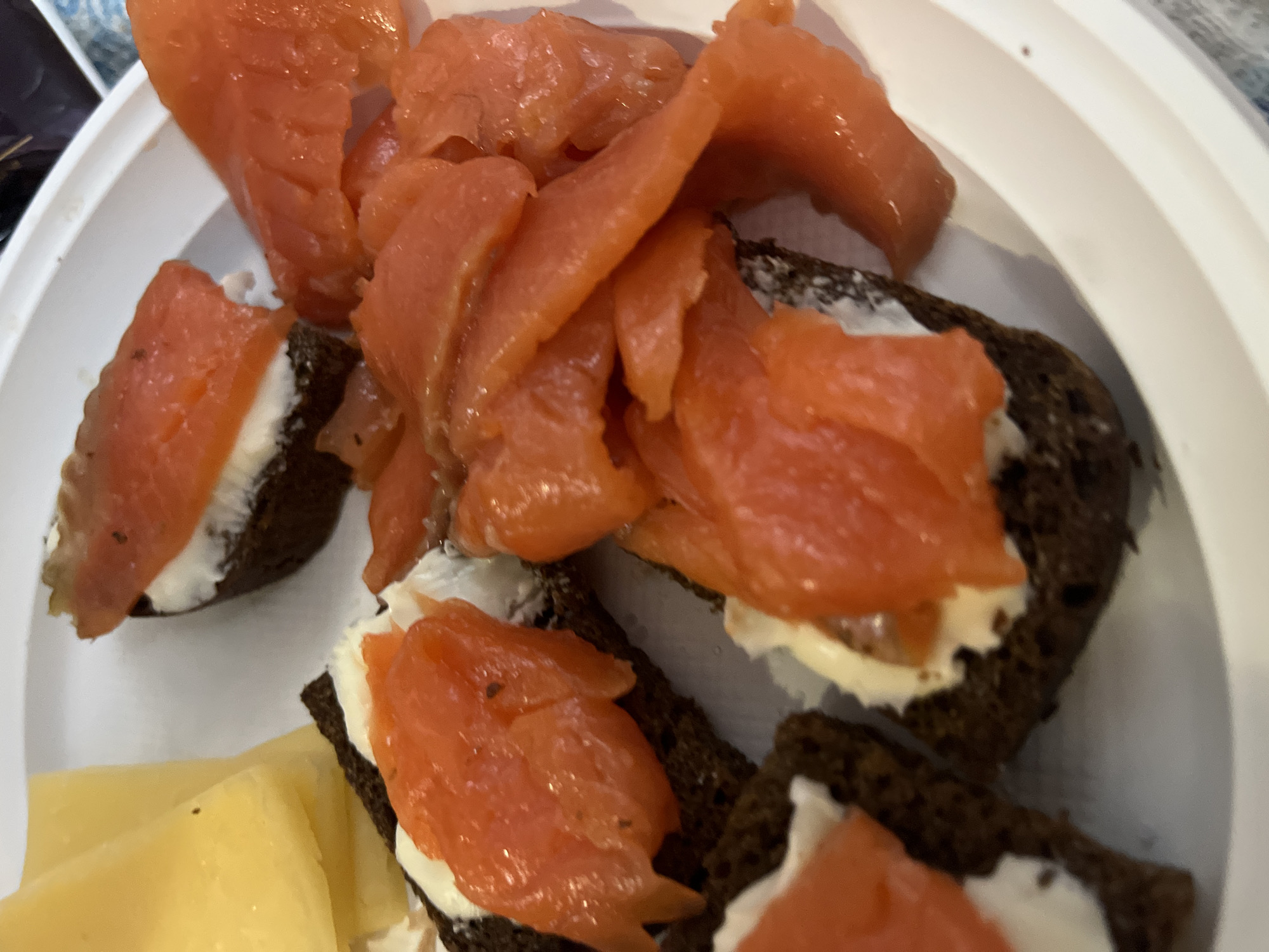 A close-up shot shows a white plate filled with rye bread sandwiches topped with a white spread (possibly cream cheese) and slices of salmon. Some salmon is also piled separately on the plate. Additionally, there are slices of yellow cheese present. The location is potentially Гаревая, Russia based on the user's request. The scene depicts a food presentation, most likely a snack or appetizer.