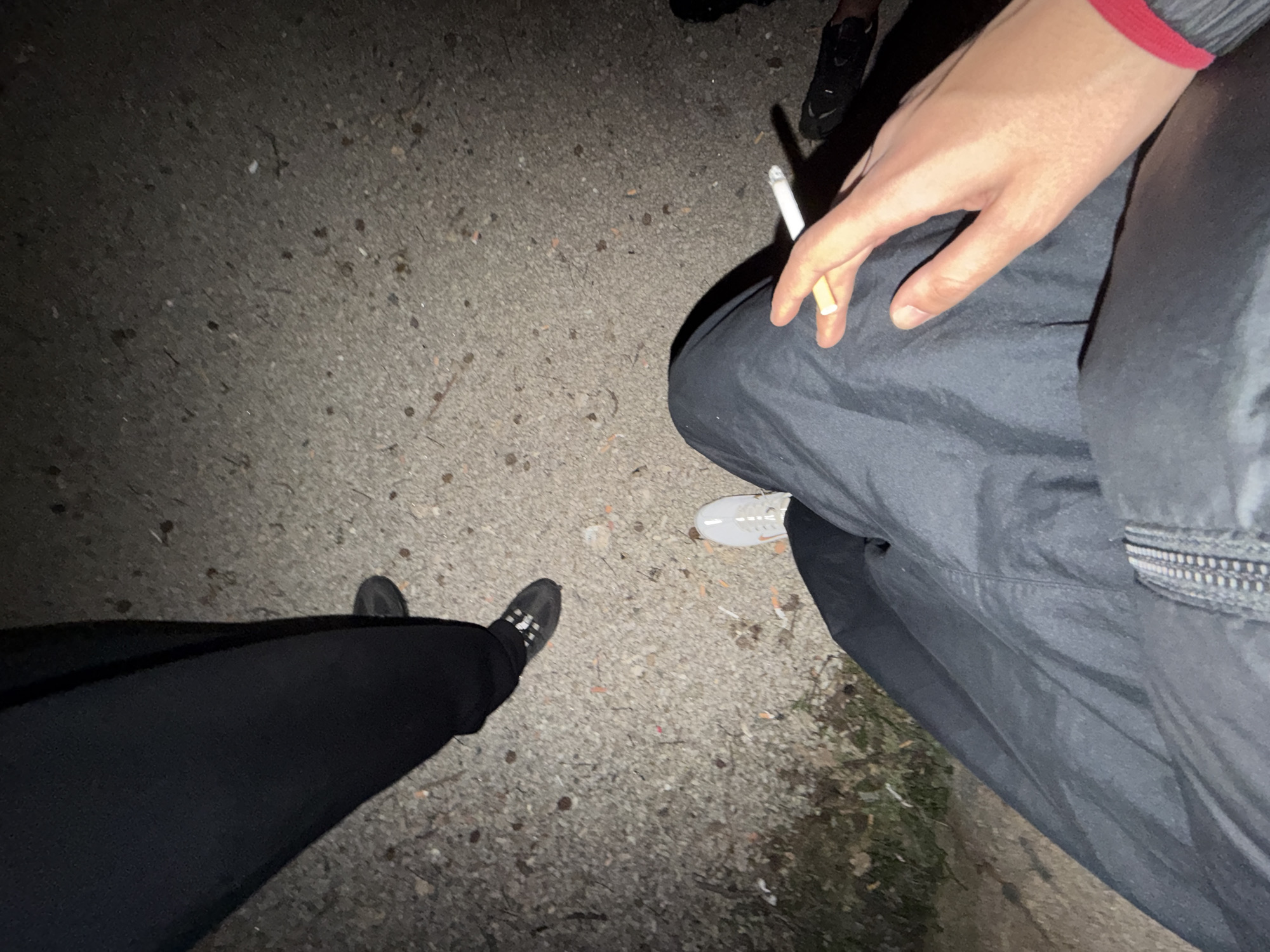 The image depicts an outdoor scene at night, likely in Vila Real, Portugal, although specific city landmarks are not visible. Three individuals are partially visible. One person, positioned on the right side of the frame, is holding a lit cigarette between their index and middle fingers. This person wears dark gray outerwear with a red wristband. Another individual is standing on the lower-left, wearing black trousers and dark shoes. A third person is partially visible at the top of the image, wearing dark-colored shoes. The setting appears to be a paved area, possibly a sidewalk or road, with debris scattered across the surface. Patches of grass or vegetation are visible near the lower right edge. The lighting is dim, suggesting the time of day and an absence of strong direct light sources.