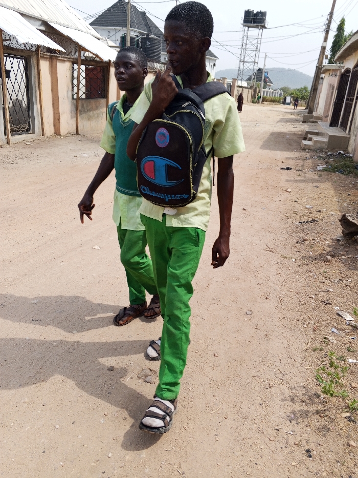 This image depicts two young males walking on an unpaved dirt road in Adonkolo, Nigeria, under clear daylight. The male in the foreground, positioned right of center, wears a light yellow short-sleeved shirt, green trousers, and white and black strap sandals. He carries a black backpack with a prominent "Champion" logo (red 'C' with blue outline) over his right shoulder and has his right hand near his mouth. The second male, positioned slightly behind and to the left, wears a similar light yellow shirt, green trousers, and a dark green sleeveless vest, with brown open-toed sandals. His left hand is raised, fingers splayed. Both individuals cast distinct shadows to their left.
The road surface is light brown dirt with scattered small rocks and sparse vegetation along the edges. In the background on the left, a light-colored building with a corrugated metal fence and gated entry is visible, followed by a building with a dark roof. On the right, another building with a light wall and dark door is present. Further down the road, a tall metal water tower with a black tank is visible, along with electrical wires traversing the scene. Distant hills or mountains line the horizon under a hazy sky. Several other figures are visible walking in the distance along the road.