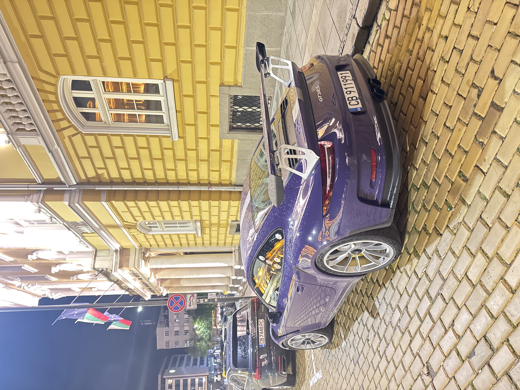 The image depicts a purple Porsche GT3 RS parked on a cobblestone street in Sofia, Bulgaria. The license plate reads "CB 911991". Adjacent to the Porsche is a black Audi with license plate "CB 7448 PC". Both cars are parked along a yellow building with distinctive brick-like facade. Overhead, the Bulgarian and European Union flags are visible. A "no stopping" sign is posted nearby. The setting appears to be in a historic or central part of the city, given the architectural style of the buildings. The scene is likely at night or dusk. The cars and street are wet, suggesting rain. The reflection in the car window shows the outline of a city street, likely filled with pedestrians.