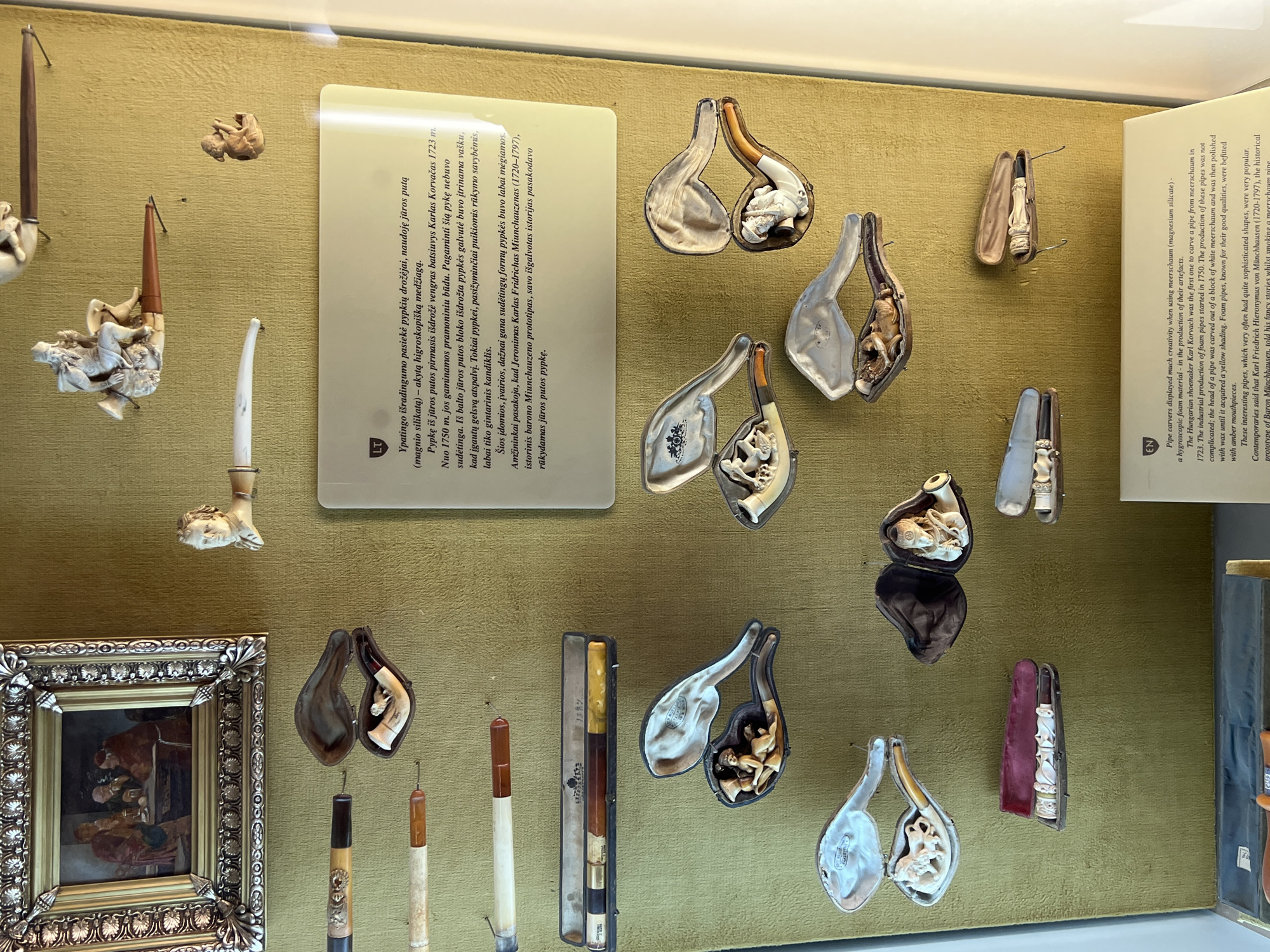 The image shows a museum display of elaborately carved meerschaum pipes, likely in the Trakai Island Castle Museum in Trakai, Lithuania. The display features numerous pipes and pipe cases arranged on a textured, olive-green wall. In the upper left corner, a small framed painting depicts a group of men gathered around a table, possibly in a tavern setting. To the right of the painting, a small, carved bust of a man's head serves as the bowl of a pipe, which extends to the right. Another small carved figure of a cherub is positioned to the right of the bust.
The majority of the display consists of various meerschaum pipes and their associated cases. Many of the pipes are intricately carved with figures, including reclining nudes, mythological scenes, and human busts. Some of the pipes are presented open within their velvet-lined cases, showcasing the detailed craftsmanship. One case in the lower center holds a pipe with multiple carved figures along its length. To the right of that, another case contains a pipe with a carved human figure at the bowl. In the upper right quadrant, a pipe with a seated figure is visible in its case. A rectangular case at the top center contains two pipes.
A descriptive plaque is mounted on the wall below the pipe carvings. The plaque has a heading with "LT" and "EN" indicating multilingual information. The English text describes the creativity in pipe carving using meerschaum, highlighting that the industrial production of foam pipes began in 1750. It mentions Karl Korvach of Hungary as one of the first to carve meerschaum pipes and notes that the pipes were polished with wax to achieve a yellow hue. The text also refers to Karl Friedrich Hieronymus von Münchhausen and his supposed stories told while smoking these pipes. The overall environment appears to be a well-lit museum interior, with the display cases and artifacts well-preserved and presented for public viewing.