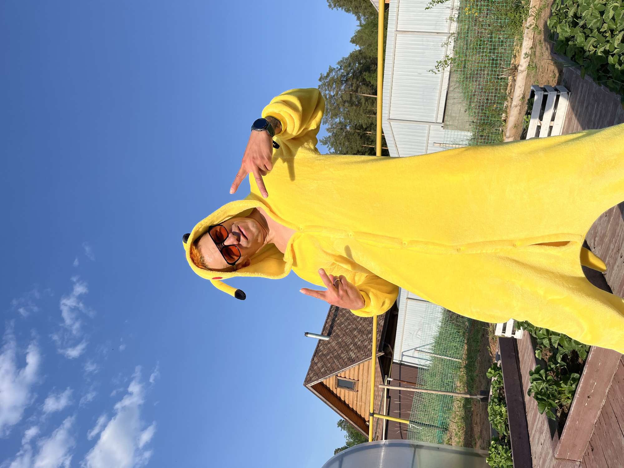 A person wearing a Pikachu costume, glasses, and a wristwatch makes a hand gesture in the direction of the camera. The environment includes a clear, cloudless blue sky, a fence, trees, a small house, a small garden, and a greenhouse. The photograph appears to have been taken in Chaikovsky, Russia.