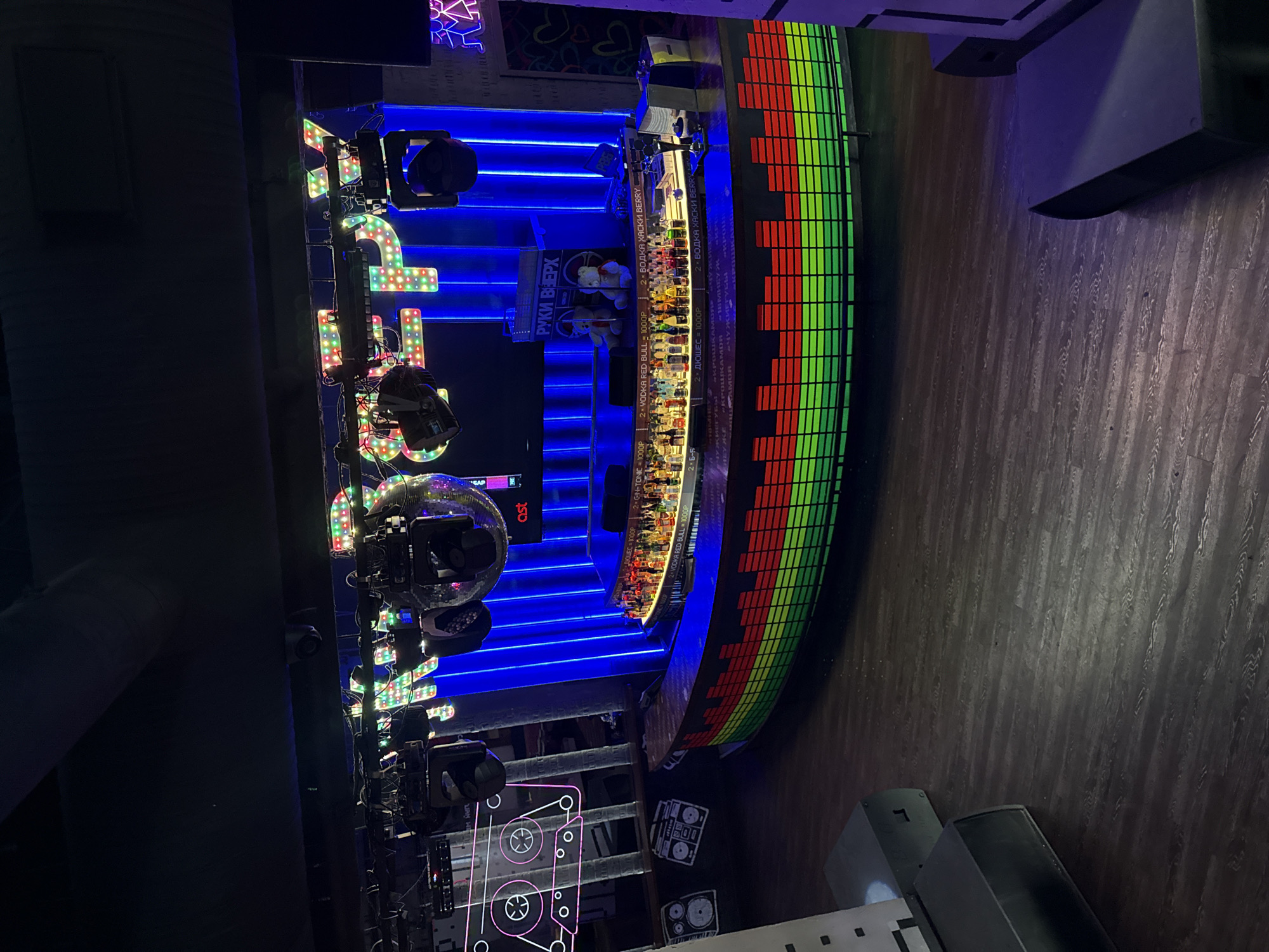 This image captures an aerial view of a bar in Moscow, Russia, featuring vibrant, dynamic lighting. The focal point of the scene is a curving bar counter illuminated with a display reminiscent of an audio equalizer, showcasing alternating red and green segments. Above the bar, an assortment of bottles is visible. The backdrop is highlighted with blue neon lighting, augmenting the atmosphere.
To the left of the bar, there are lighting fixtures, including mirror balls and stage lights. A sign reading "РУКИ ВВЕРХ" can be seen. The ground is covered with a wooden flooring pattern. A cassette tape design is visible, adding a vintage aesthetic to the environment.
The scene is devoid of people, highlighting the setting rather than the patrons of the bar.