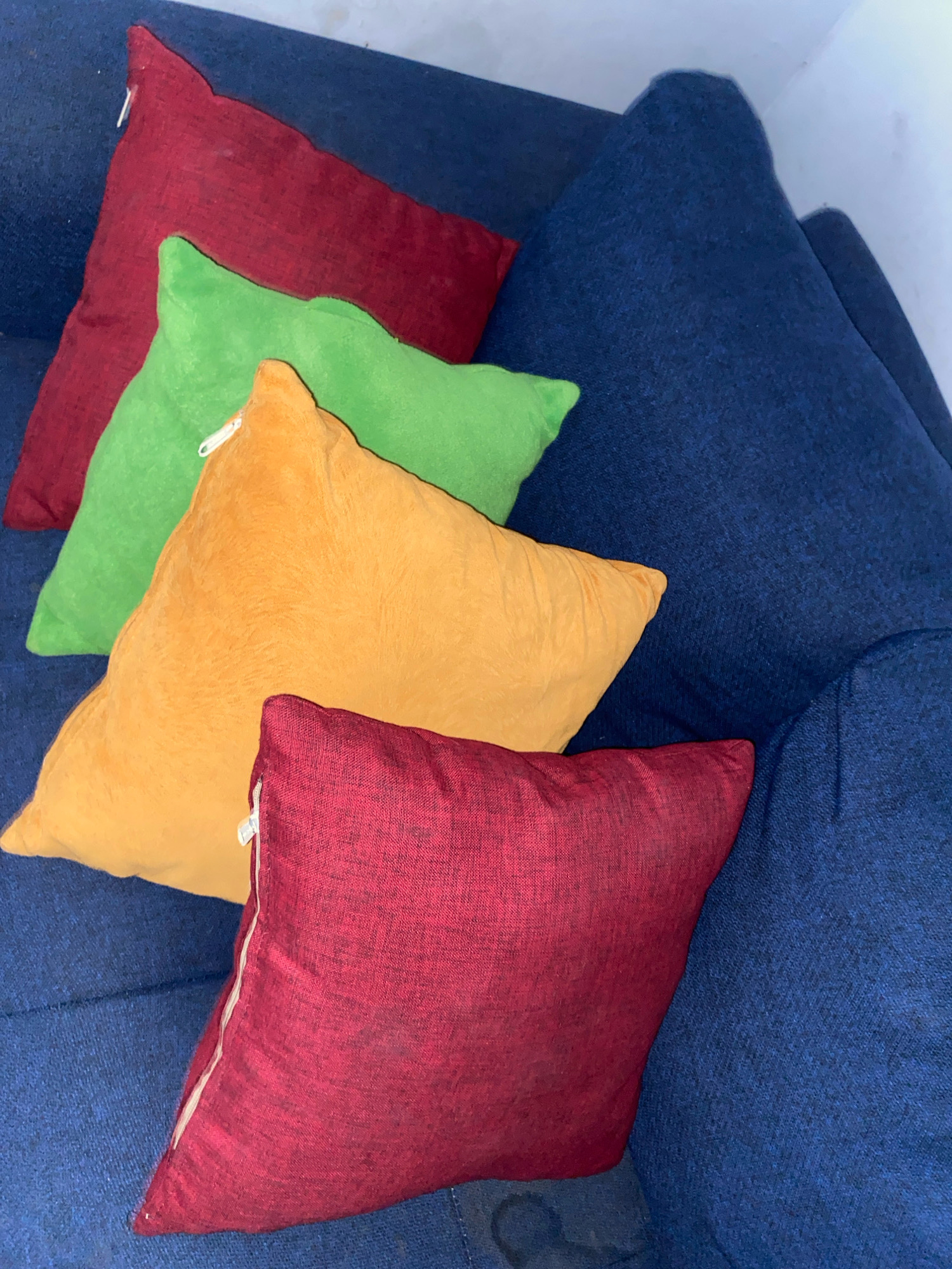 A collection of four square throw pillows is arranged diagonally on a blue upholstered sofa. From the top left to bottom right, the pillows are colored deep red, bright green, orange, and a darker shade of red. The sofa is a dark blue, textured fabric. The image appears to be an indoor setting, likely a living room or den, with a portion of a white wall visible in the upper right corner. There are no people or animals present in the scene. The location in Klurak, Indonesia is not discernible from the visual information provided in the image.