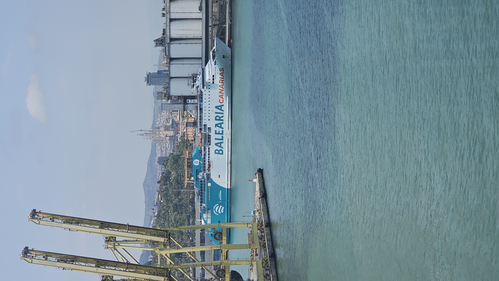 A daytime shot depicts a ferry docked at the Port of Barcelona, Spain. The ship is emblazoned with "BALEÀRIA CANARIAS" in large, vertically-aligned lettering. Dockside infrastructure, including tall yellow cranes, is visible in the foreground on the left. A pier extends into the light-green-colored water. In the background, the city's skyline, including its notable structures such as the Sagrada Familia, rises inland from the port. The sky is overcast.