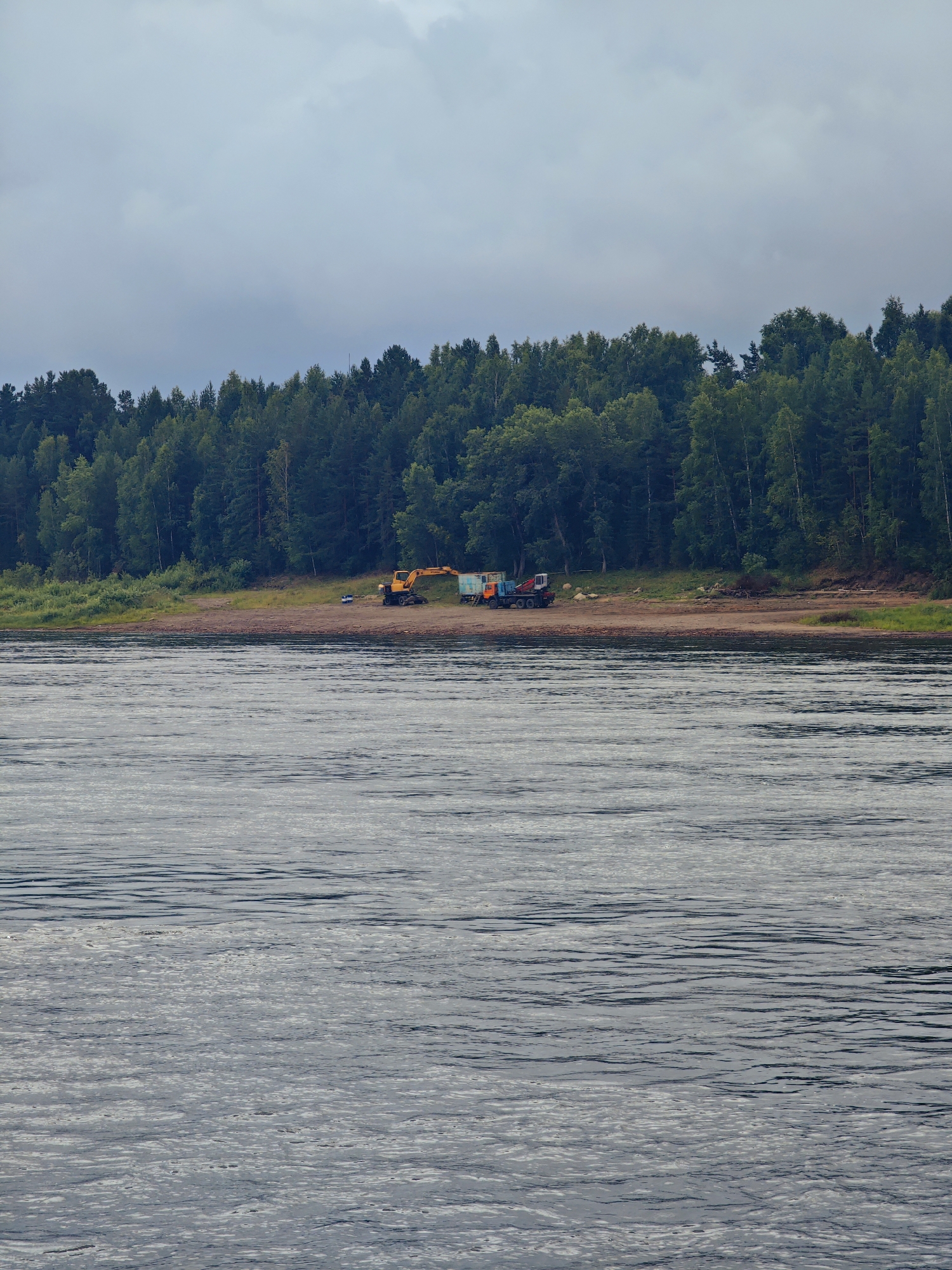An aerial shot captures a scene on the Yenisei River bank in Lesosibirsk, Russia. The river occupies the foreground, its surface rippled by a gentle current. In the middle ground, a sandy shore is visible, lined with a dense forest of coniferous trees that form a dark green backdrop. On the shore, two construction vehicles are parked: a yellow excavator and a blue and orange truck with a crane. The sky above is overcast with a pale grey hue, suggesting a cloudy day. The overall impression is one of a remote, natural landscape with signs of industrial activity.