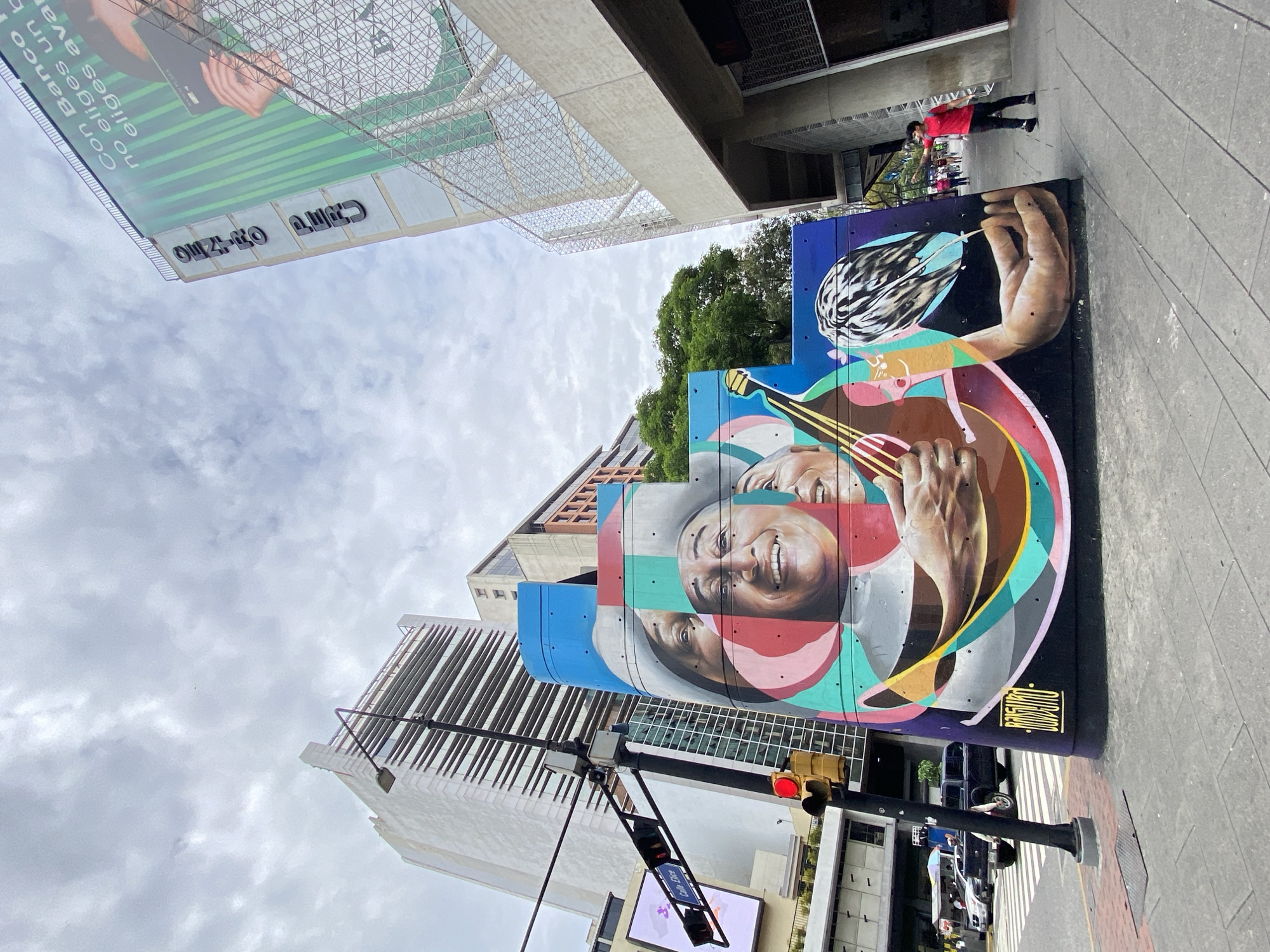 This full-color, upright street photograph captures a vibrant urban scene in Caracas, Venezuela, under a cloudy sky. The primary subject is a large, colorful mural painted on a prominent structure on the right side of the frame.
The mural depicts a man with a wide, joyful smile, his face rendered in a fragmented, geometric style using bright colors like red, blue, green, and brown. He is shown playing a stringed instrument, possibly a mandolin or cuatro, with a detailed hand strumming the strings. A signature, "ROBOGUITO," is visible at the bottom right of the mural.
The background reveals a bustling city environment with tall, modern buildings featuring glass and concrete facades. On the left, a large advertising billboard is supported by a metal framework. The billboard displays an advertisement with a person's hand holding a phone against a green and white striped background. Visible text on the billboard, when correctly oriented, includes "Con Banco..." and large black letters spelling "GUZMAN" and "CITI."
A sidewalk runs along the foreground and right side, where a person in a red shirt and dark pants is visible. A traffic light pole with a red signal and a street sign (partially obscured, showing "Calle...") mark an intersection. Several cars are partially visible on the street. The scene is illuminated by diffused daylight, typical of an overcast day, portraying a lively urban landscape infused with public art.