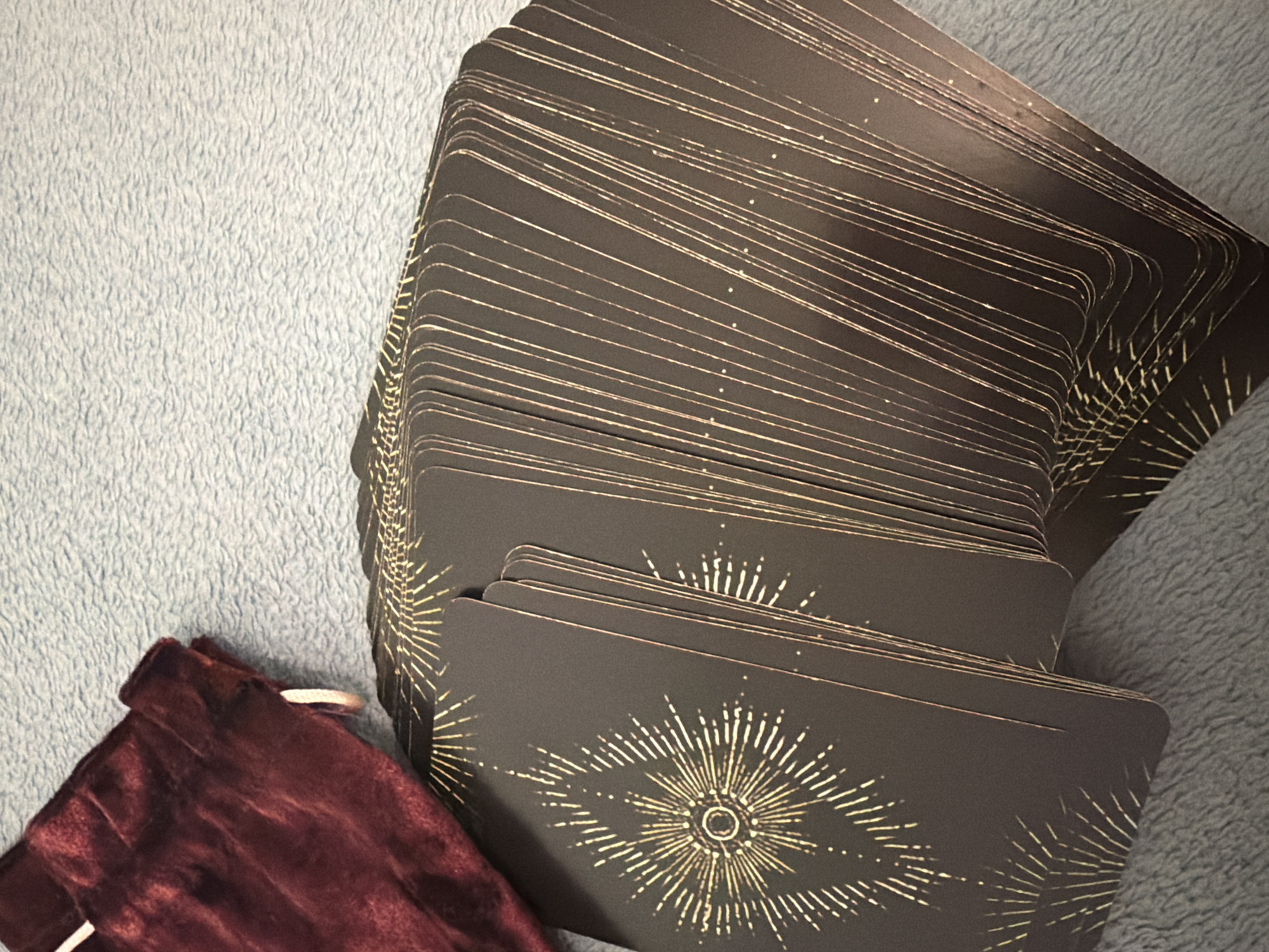 The image shows a fanned-out deck of tarot cards with a gold sunburst pattern on a dark background. To the left of the cards is a burgundy velvet drawstring bag. The objects are resting on a light blue textured surface, likely a blanket or rug. The scene is depicted from a high-angle, close-up perspective, with the lighting casting subtle shadows. There are no people or actions visible in the image. The location is not specified in the image and there is no indication of Sharm El Sheikh, Egypt.