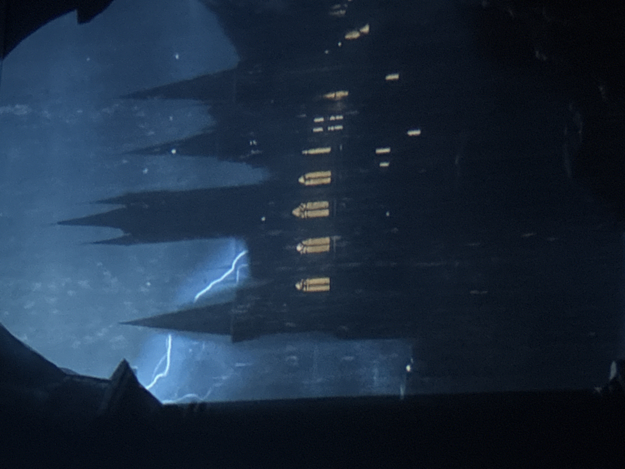 The image shows a nighttime view of a tall, dark structure with multiple pointed peaks, resembling a castle or gothic-style building. The structure is illuminated by internal lights that appear as yellow rectangles arranged vertically. In the foreground, there is an out-of-focus dark shape that could be part of a window frame or another structure. In the background, lightning flashes illuminate a cloudy sky. No people are visible. The location is not discernible as Varna, Bulgaria solely from this image. The scene depicts a dramatic, possibly ominous, atmosphere.