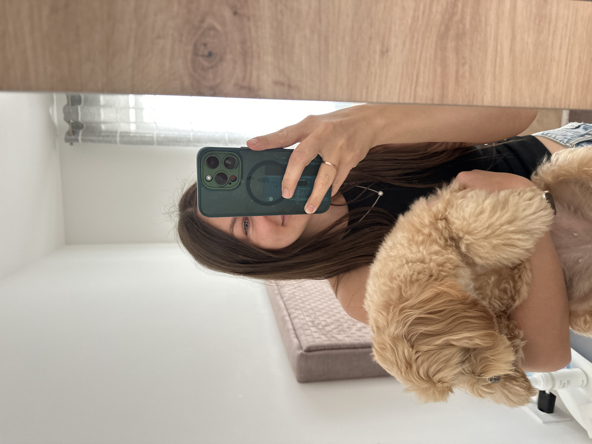 A woman is taking a selfie with a dog in front of a mirror. The woman has long brown hair, blue eyes, and is wearing a ring. She is holding a phone with a green case. The dog is small, with tan curly fur. The background includes a wood-grain surface, a window with light curtains, and a wall. The location is Donji Pobori, Montenegro. A bed or cushion is visible on the floor.