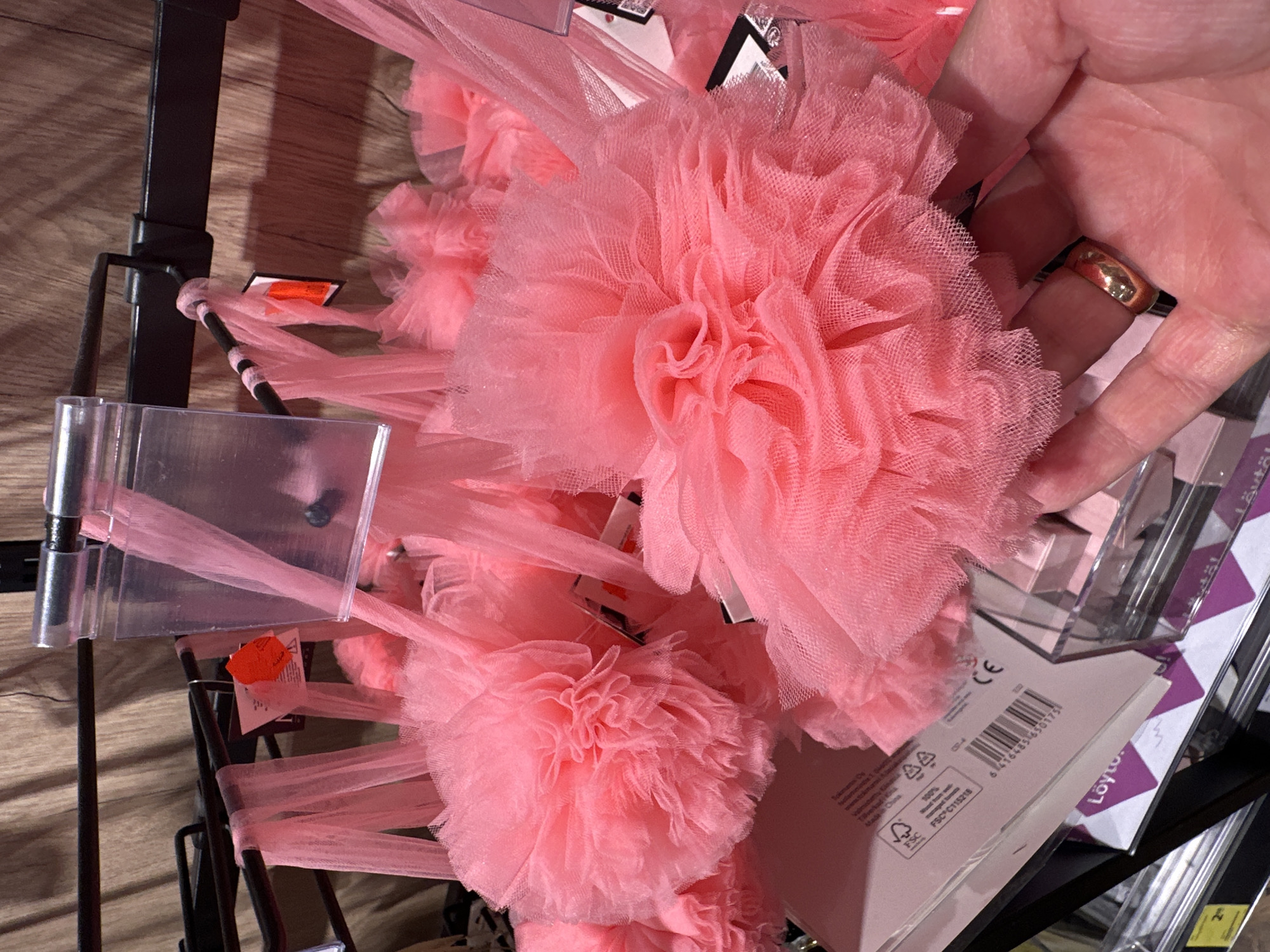 The image shows a close-up view of several bright pink, fluffy tulle pom-poms hanging on a display rack in what appears to be a retail store. The pom-poms are the primary focus, with their intricate layers of sheer fabric creating a soft, voluminous texture. They are suspended by thin pink straps. Behind the pom-poms, a wooden-paneled wall is visible, along with parts of a black metal display structure.
In the foreground, a hand is gently touching one of the pom-poms. The hand has a ring on the ring finger, which is a simple gold band. The hand is positioned to the right side of the frame, partially obscuring some of the pom-poms.
On the left side of the image, part of a pink retail box or packaging is visible, with text and a barcode printed on it. This packaging also has a small green FSC logo, indicating it is made from responsibly sourced materials.
The lighting in the image is bright, highlighting the vibrant pink color of the pom-poms. The overall impression is one of a retail display for decorative or novelty items. The location is described as "Unknown, Unknown," indicating that the specific city and country are not provided.